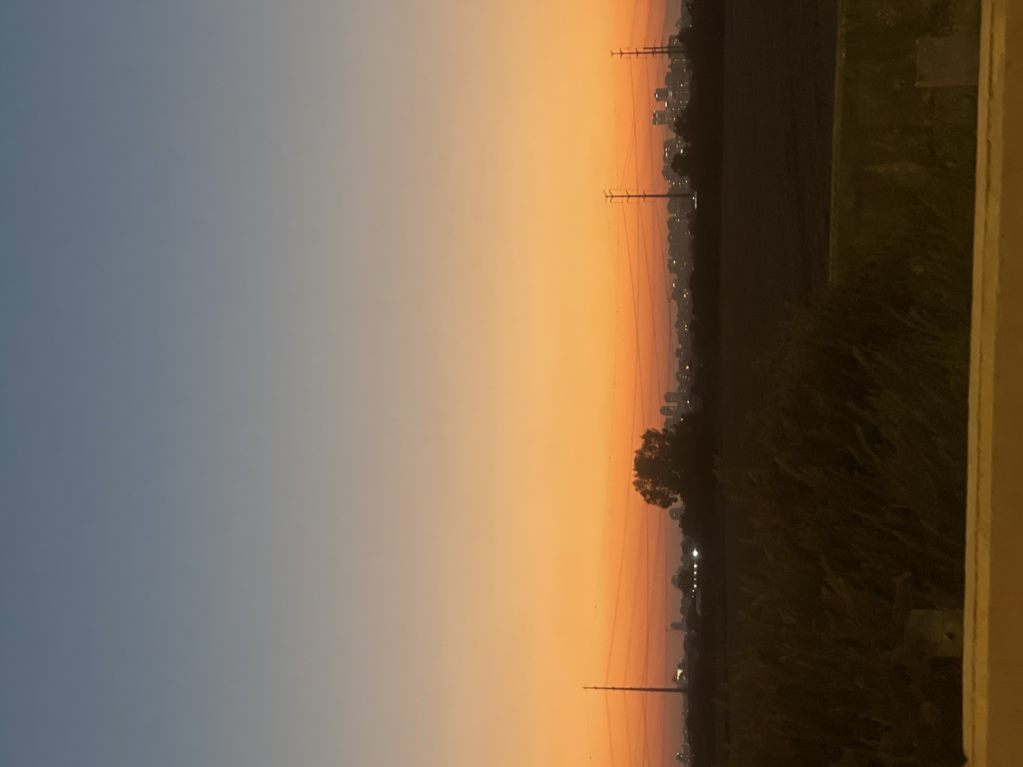 The image depicts a landscape at dusk with a city skyline in the distance. The sky is a gradient of blue and orange, transitioning from a light blue at the top to a vibrant orange near the horizon. In the middle ground, there are fields and some vegetation. Two tall power line towers are visible, with power lines stretching between them. Silhouetted against the orange sky are buildings and trees, suggesting a populated area. The foreground shows a blurry yellow barrier or railing, and dark, dense vegetation. The overall impression is that of a serene, urban fringe at sunset. The location is specified as Unknown, Israel.