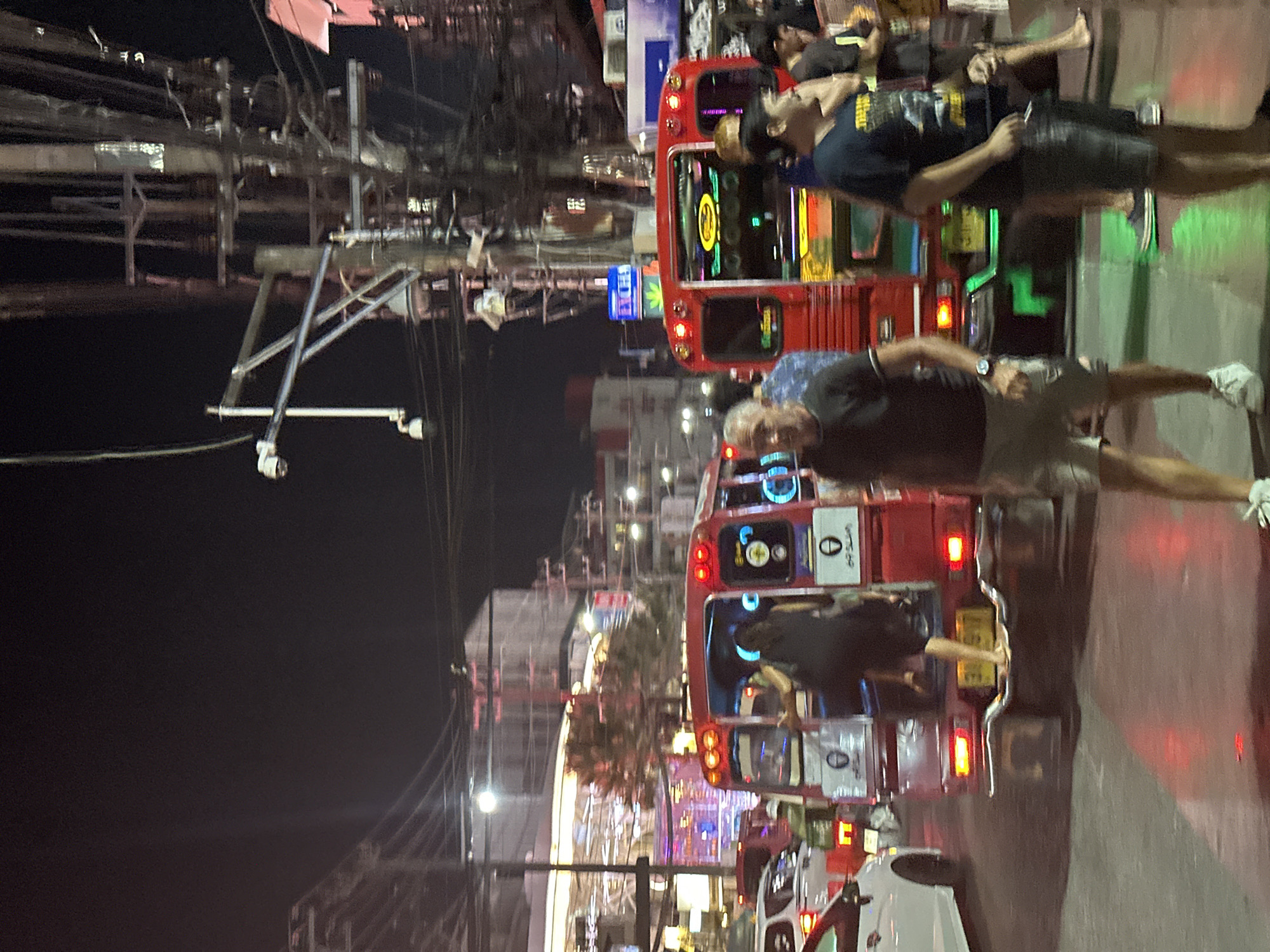 The image depicts a nighttime street scene in Patong, Thailand. The environment is characterized by dense overhead utility wiring and artificial illumination from various sources.
The scene focuses on a multi-lane street occupied by both vehicles and pedestrians. Two red, open-air passenger vehicles, typical of "songthaews," are prominent in the midground. The leftmost red vehicle shows a female individual in a dark dress in the process of boarding its rear passenger compartment, her left foot elevated onto a step and her right foot bare. This vehicle's rear features two red taillights, illuminated brake lights, and a yellow Thai license plate (partially legible). The rightmost red vehicle displays its rear interior illuminated by multicolored neon lights and two red taillights. Above this vehicle, a blue "EXCHANGE" sign with a green cannabis leaf symbol is visible.
In the foreground, a male with short grey hair, wearing a dark t-shirt, light shorts, and white athletic shoes, walks towards the viewer, his left wrist displaying a silver-colored watch. To his immediate right, another male, wearing a dark t-shirt and dark shorts with dark footwear, also walks towards the viewer. Additional individuals, some blurred, are present in the background and near the right red vehicle; one person near the right vehicle appears barefoot.
The road surface exhibits reflections of red and green light, indicative of moisture or a reflective material. A white sedan is partially visible on the far left of the frame, positioned in front of the leftmost red vehicle.
On the right side of the image, a utility pole is heavily laden with numerous black electrical and communication cables. A metal cross-arm extends horizontally from this pole, supporting at least two white, dome-shaped CCTV cameras. The background features multi-story buildings, illuminated with white and colored lights, typical of urban commercial establishments. The sky is dark, confirming a nighttime setting.