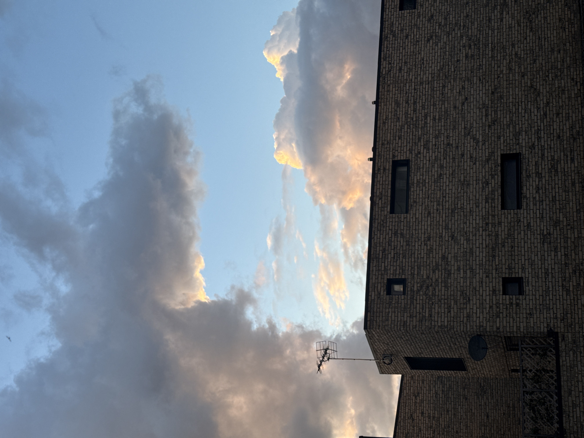 The image shows a building facade and sky with clouds. The building is made of dark brick, with a rectangular grid pattern. It has several small, rectangular windows and a satellite dish. An antenna extends from the building. The sky is partially obscured by thick, dark clouds, some tinged with yellow. There is a patch of blue sky visible. The photo appears to be taken in the evening or early morning based on the lighting. There is no other information.