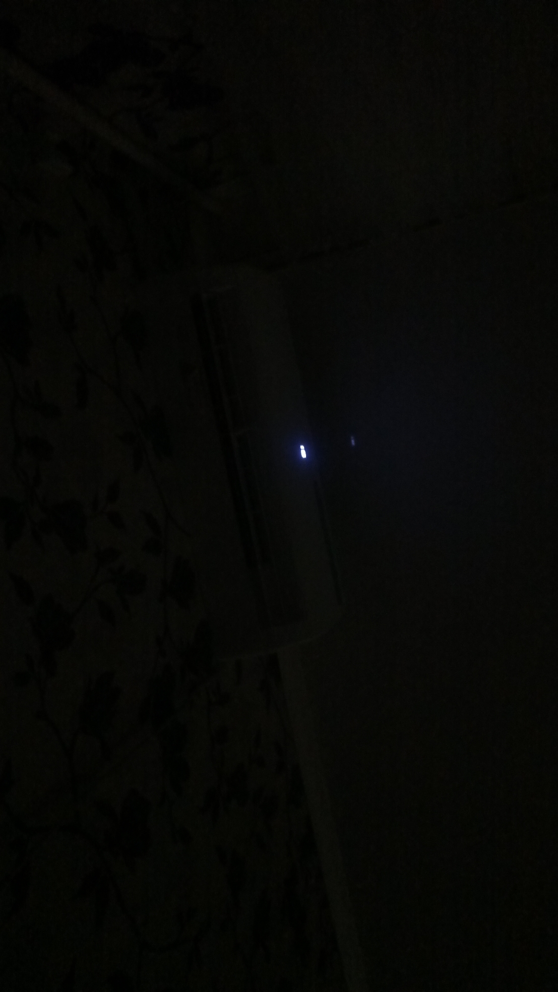 The image depicts a dimly lit interior space. On the left, a wall is covered in dark, floral-patterned wallpaper. To the right, a white, rectangular object with a glowing blue light on its surface is visible. This object appears to be an electronic device, possibly a router or a modem, positioned against a darker background. The overall ambiance is dark, with minimal illumination. No people or specific city details are discernible from the image.