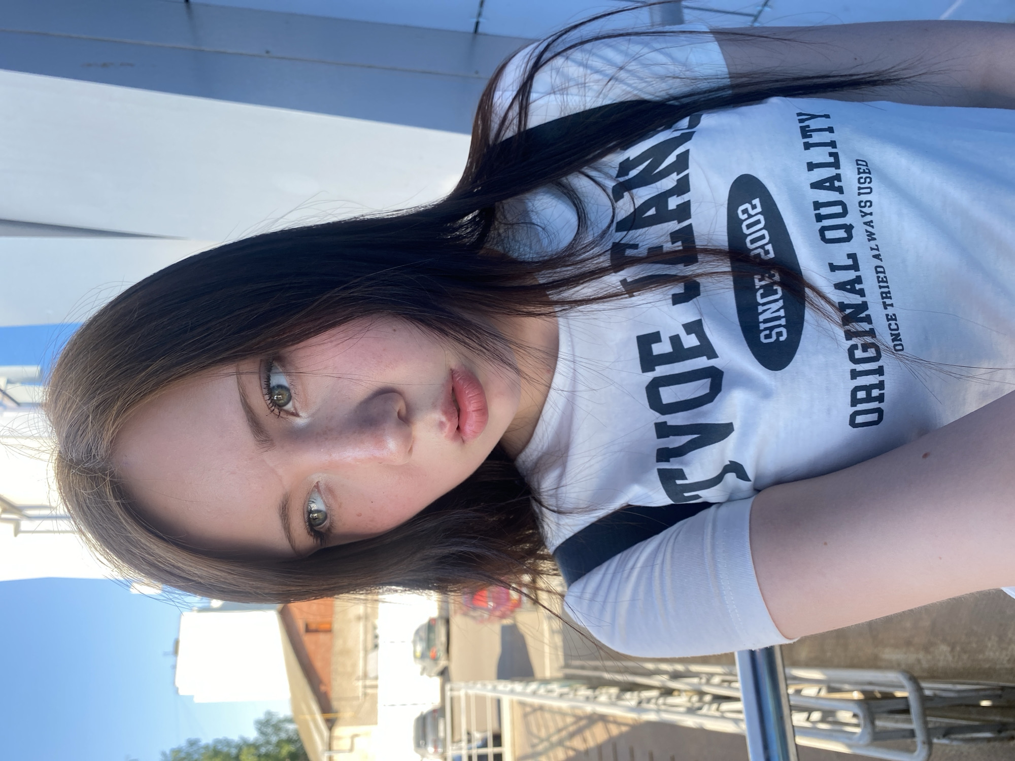 A woman is looking into the camera in an outdoor setting. She has fair skin, dark brown hair with light brown highlights, and green eyes. She is wearing a white t-shirt with black writing, possibly a logo for "Tyoe Jeans" and "Original Quality." The background appears to be an urban landscape with buildings, vehicles, and possibly a bridge railing visible. The image is taken from a slightly elevated angle, suggesting the woman is looking down at the camera.
