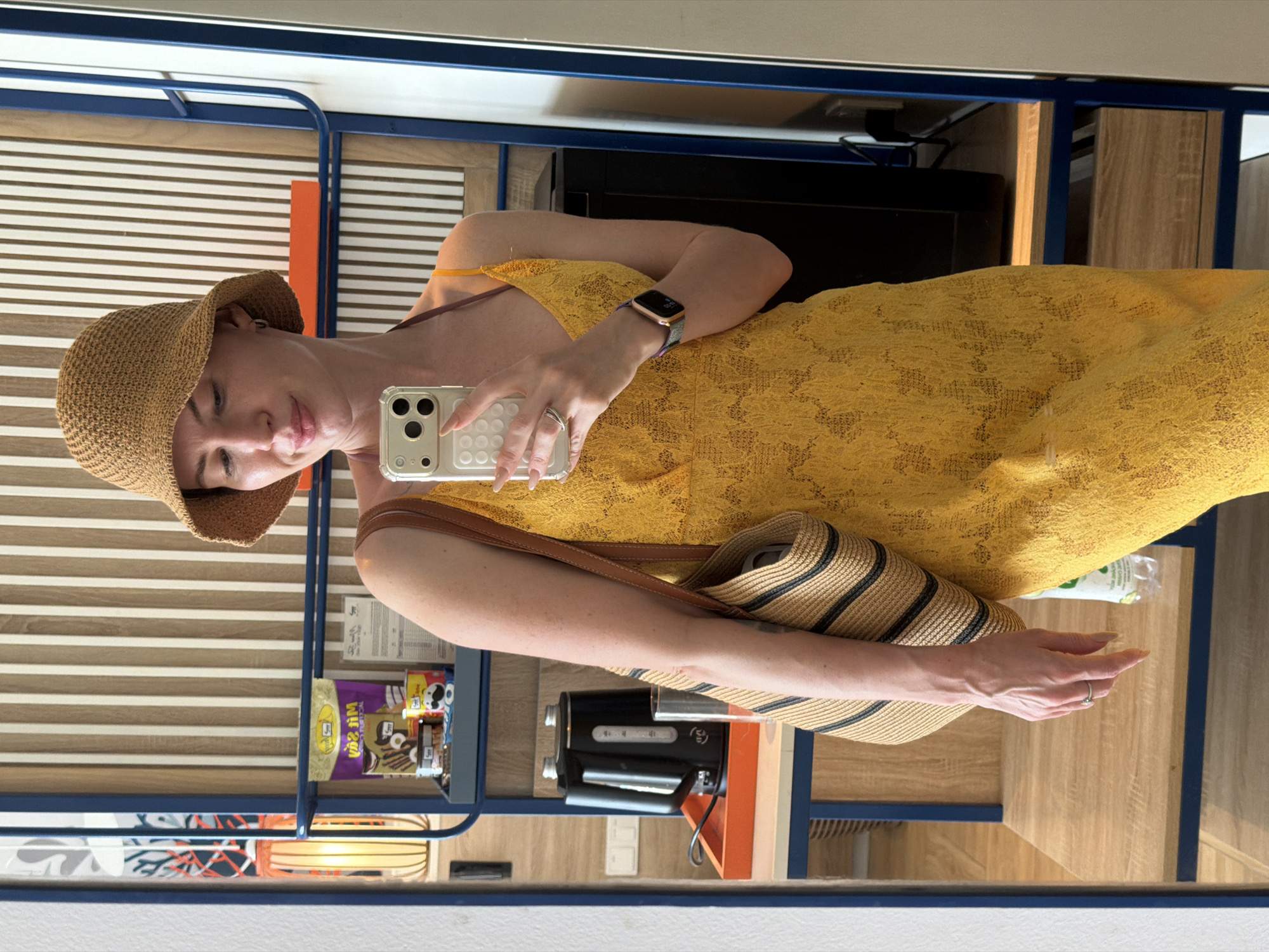 A woman stands in a hotel room, taking a selfie in a mirror. She is wearing a yellow lace dress, a straw bucket hat, and a woven purse slung over her shoulder. She is holding a smartphone in her right hand and wearing a watch on her left wrist. The room features a black electric kettle and a shelf stocked with various items, including snacks and a water bottle. The background shows striped wooden paneling.