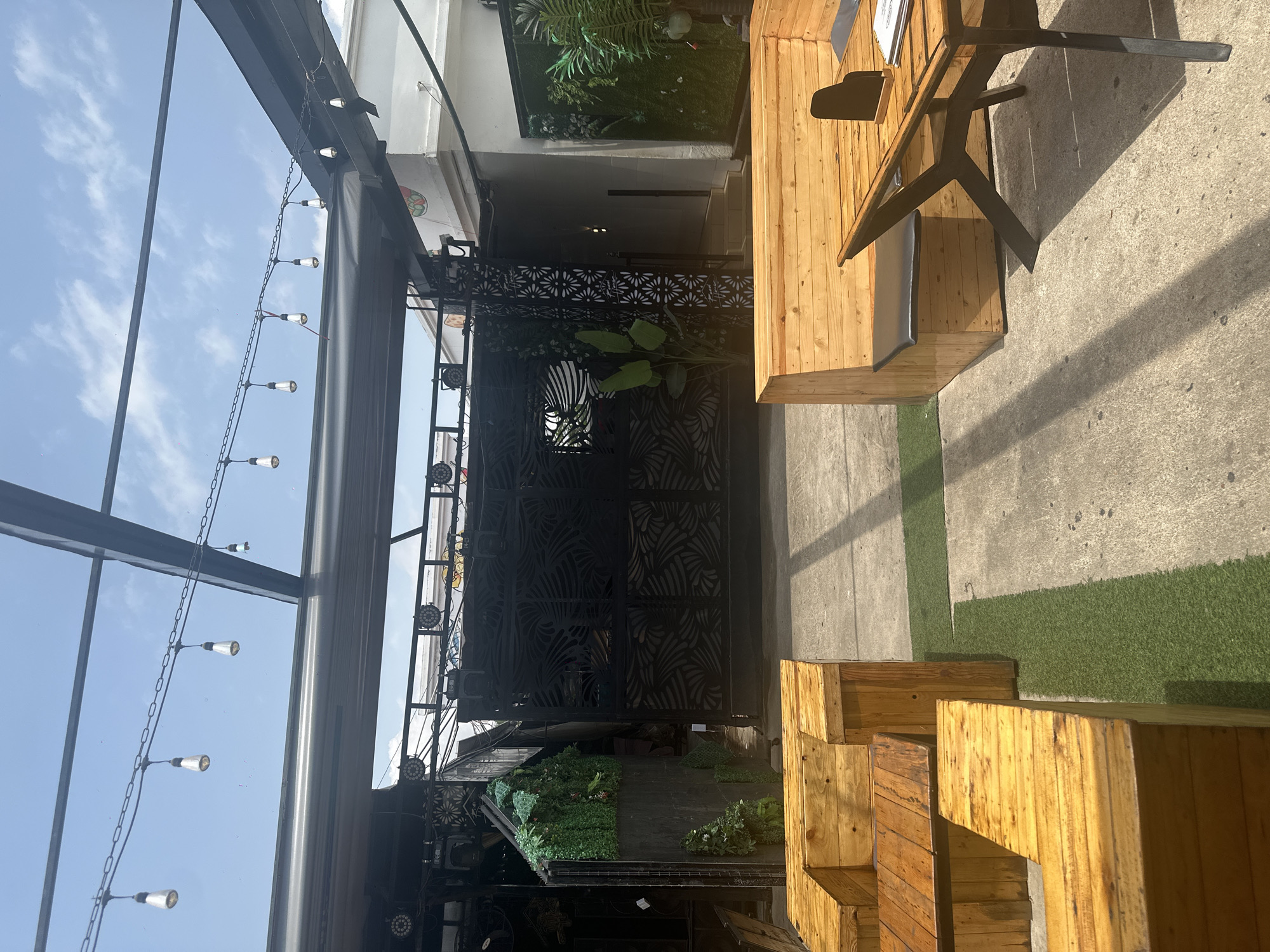 The image depicts an outdoor seating area, likely a part of a restaurant or bar, under a covered structure. In the foreground, there are several modular wooden seating units and a wooden table with a black base. The seating units appear to be made of stacked wooden planks, some with black cushions. Artificial turf is visible between some of the seating areas.
In the background, a large black metal gate with a decorative cutout pattern is prominent. To the left of the gate, there is a vertical structure covered with artificial green foliage. Above the seating area, strings of Edison-style light bulbs are suspended from a metal framework, suggesting the space is also used in the evening. Lights, possibly stage lights or architectural lighting, are mounted on structures above the gate and foliage. The environment appears to be an urban setting, possibly in Howayi, Nigeria, given the presence of tropical plants and the overall architectural style. The sky is bright blue with scattered clouds, indicating daytime. No people are visible in the scene.
