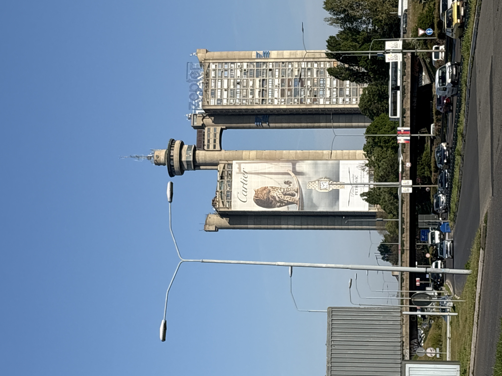 The image features the iconic Western City Gate (Genex Tower) in Belgrade, Serbia. It comprises two towers connected by a two-story bridge at the top. One tower displays a large Cartier advertisement, while the other shows the "Genex" signage. Streetlights line the adjacent road, where vehicle traffic is visible. The sky is clear blue.