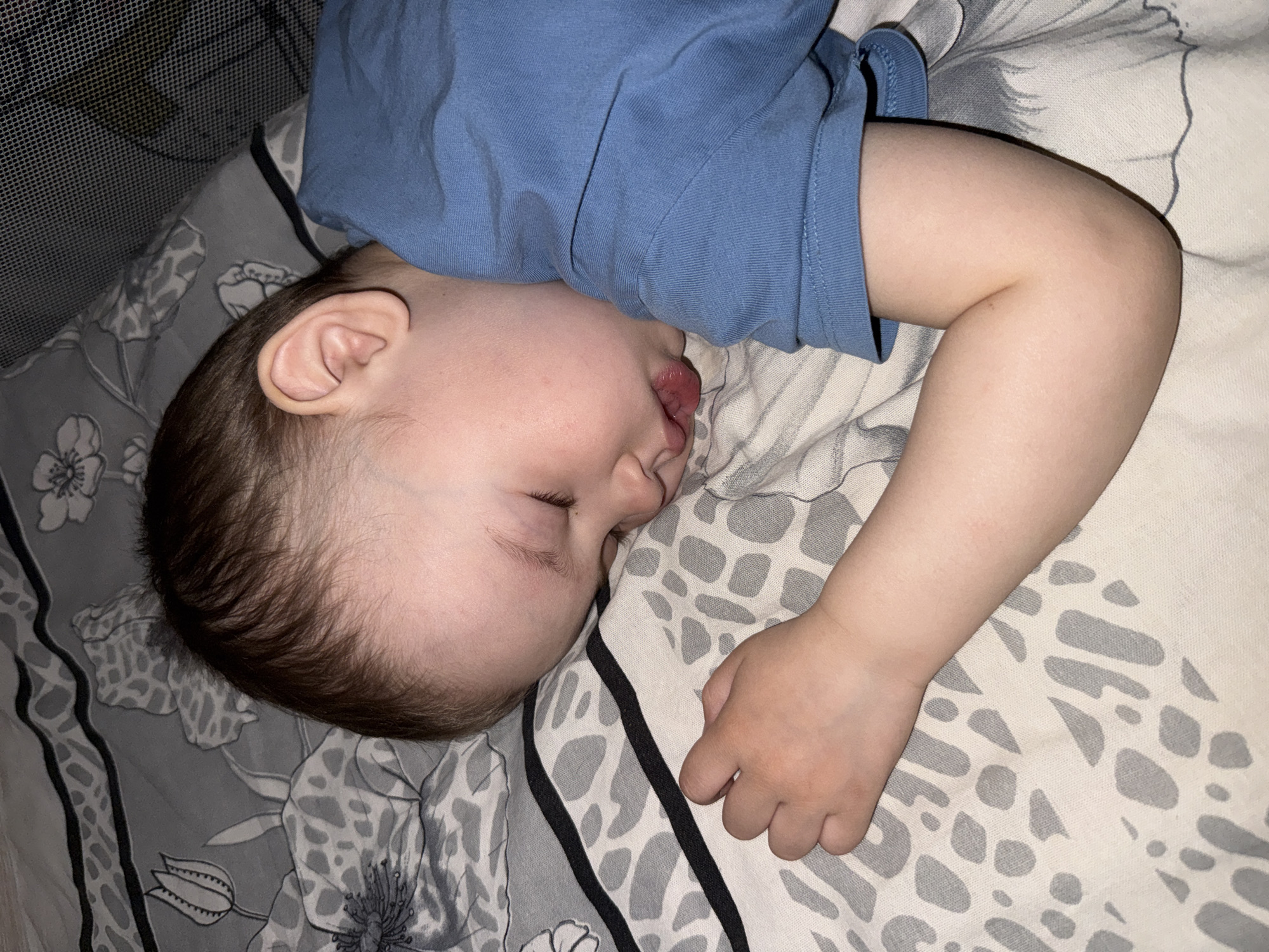 A toddler is asleep on a bed. The child, dressed in a blue shirt, is lying on their side, with their right arm extended out in front of them. The bedding is a patterned grey and white. The child appears to be resting comfortably. The photo does not contain verifiable evidence of the location within Yaroslavl', Russia.