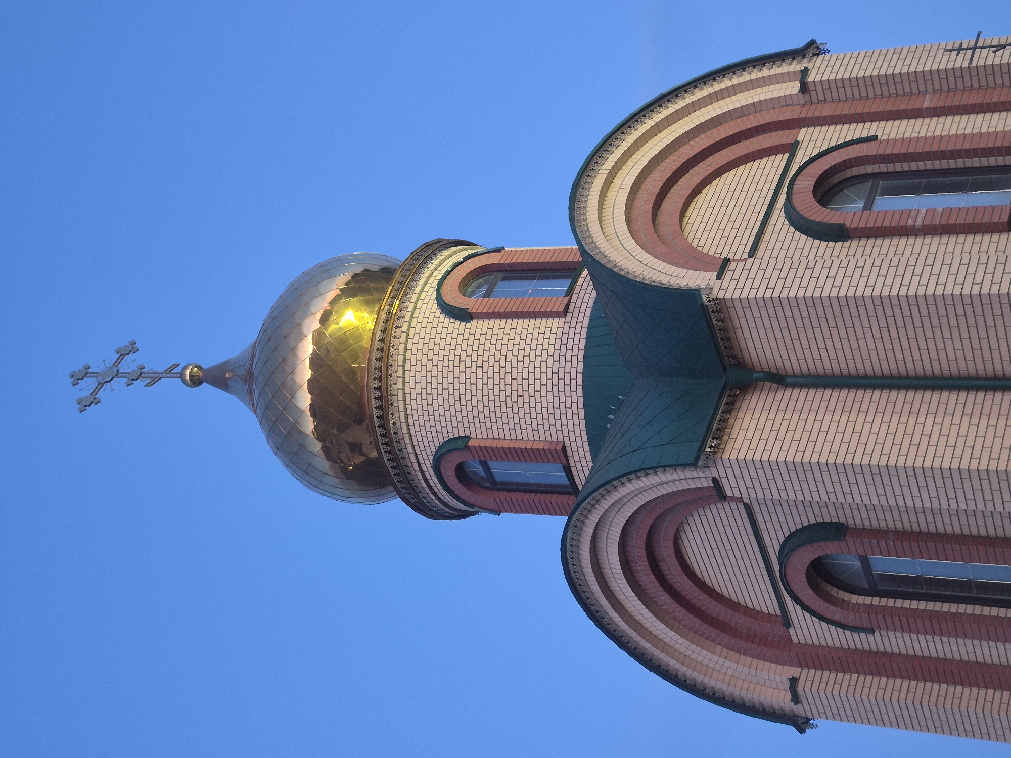 The image shows a low-angle view of a church structure against a clear blue sky. The building features a golden, onion-shaped dome topped with a cross. The dome appears to be made of metal plates, possibly copper. Below the dome is a cylindrical section, constructed of light-colored bricks. Below that is a section of building covered with curved segments of alternating shades of brick with small rectangular windows. Based on the provided information, this building is located in the city of Sergeevka, Russia.