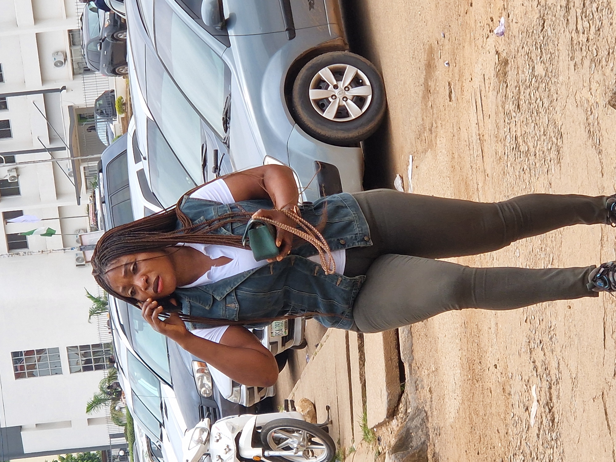 In Jos, Nigeria, a woman with braided hair, wearing a denim vest over a white polo shirt and olive green pants, walks across a dirt and gravel area. She is holding a small, dark green purse with a gold chain strap and is speaking on a black mobile phone held to her right ear. Several cars, including a silver sedan, a light blue sedan, and a dark SUV, are parked in the background. A white scooter is visible to the left of the woman. A building with a white facade and multiple windows stands behind the parked vehicles. The ground is uneven with patches of dry grass and visible debris.