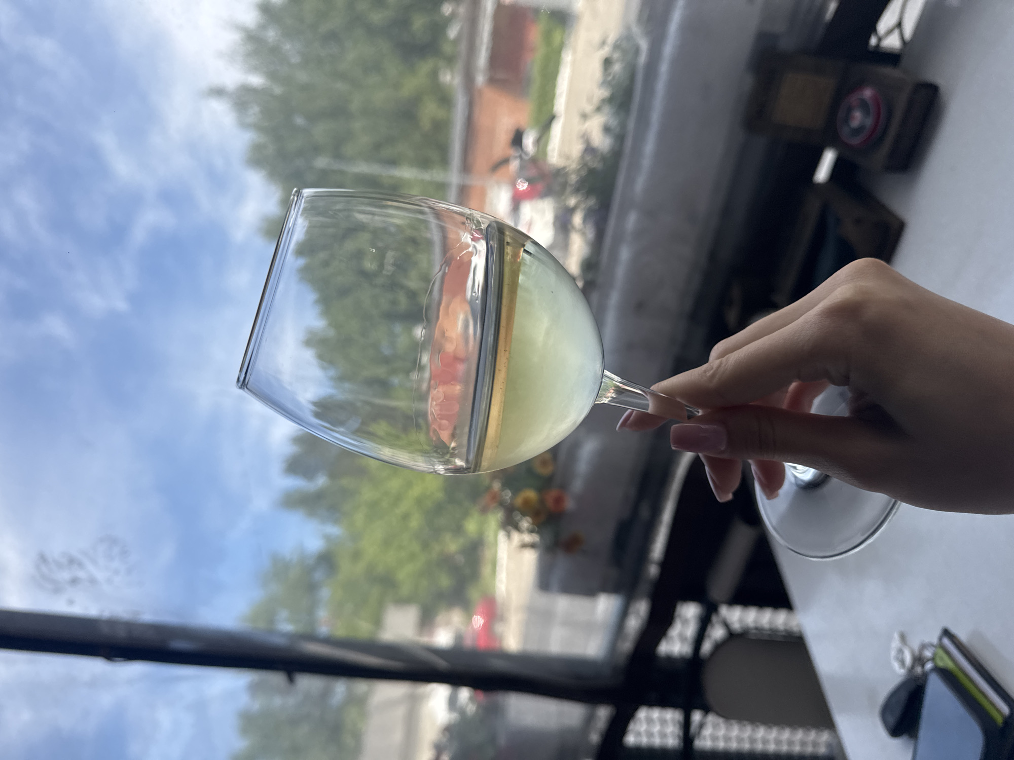 A close-up, slightly tilted photograph captures a person's hand holding a stemmed glass filled with a pale, yellowish-green liquid. The hand, with light pink polished nails, delicately grasps the stem of the glass. The scene is set indoors, looking out through a large window, suggesting a cafe, restaurant, or home in Chaikovsky, Russia.
The liquid in the glass reflects the bright, cloudy sky and green trees visible outside, indicating it is daytime. Beyond the glass, the outdoor environment shows a blue sky with soft white clouds, lush green foliage from trees, and hints of buildings. A blurred red object, possibly a vehicle or part of a distant building, is visible through the window.
On the table surface beside the hand, partially visible, are a pair of dark sunglasses and a smartphone with a distinctive bright green or yellow case. The overall impression is one of a relaxed daytime moment, perhaps enjoying a drink while observing the outside world. No discernible text is visible on any objects within the frame.