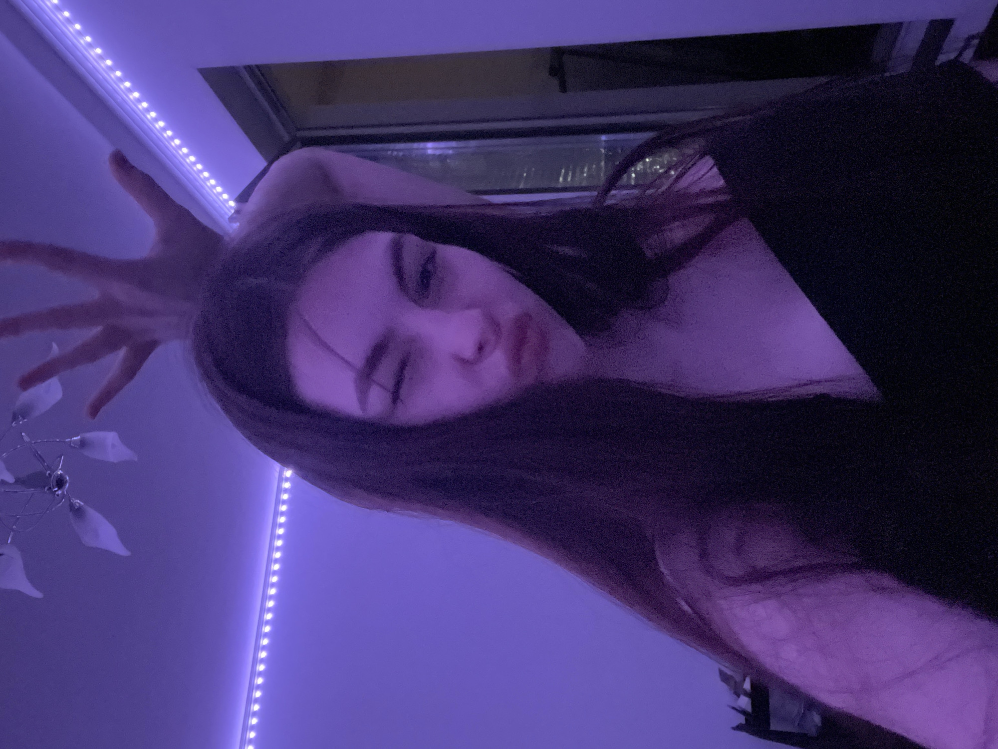 A woman is lying down in a room with purple lighting. Her long, dark hair is partially obscuring her face, but it is visible that she is winking and pouting. Her left hand is raised near her head with fingers spread. A string of LED lights is on the wall behind her, and a chandelier is also visible in the background. A window is behind her, and city lights can be seen through it, suggesting a nighttime view. No information is available to confirm the city of Saint Petersburg, Russia.