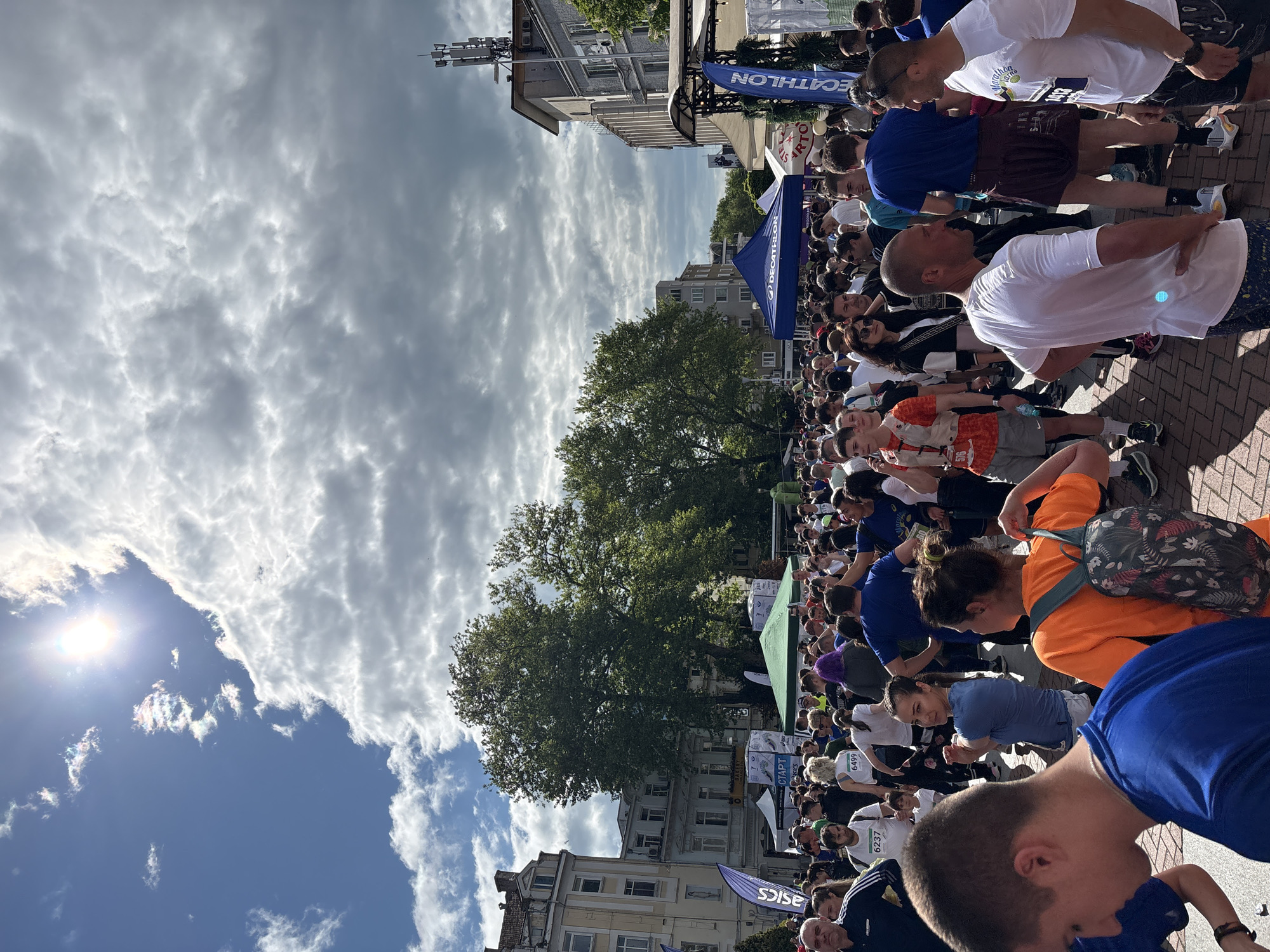 The image shows a crowd of people gathered on a street in Varna, Bulgaria. Many appear to be runners, possibly participating in a race, and are wearing bibs with numbers. The crowd is densely packed, with some people stretching or resting.
In the background are buildings and trees, typical of a city street scene. Banners and tents are set up, possibly for event support or sponsorship, including banners for Decathlon and Asics. The sky is partly cloudy with the sun shining brightly in the corner. The brick pavement suggests an urban setting.