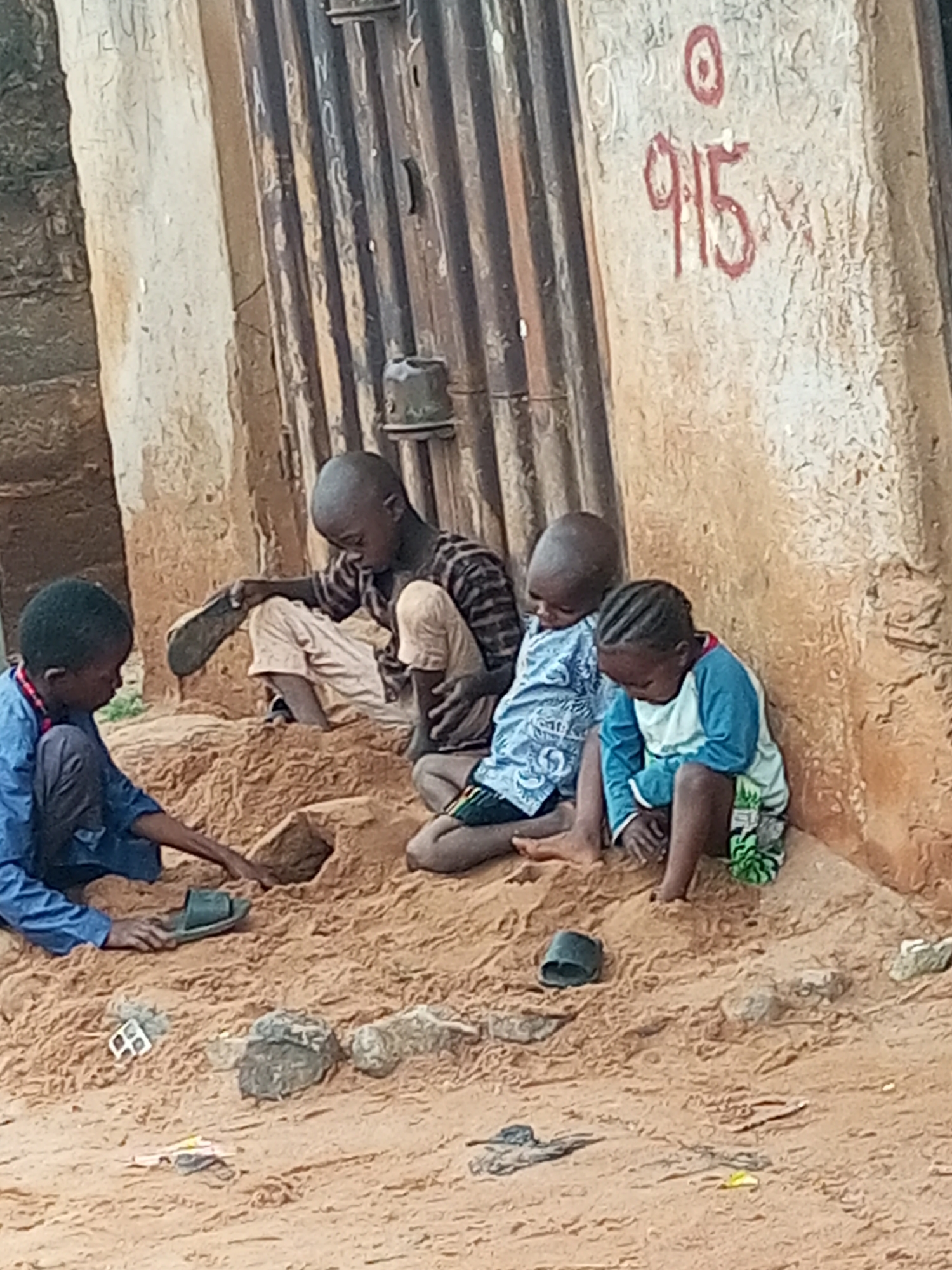 In Tumfure, Nigeria, four young boys are observed sitting in a pile of sand near a corrugated metal wall. The boys are engaged in playing with the sand. One boy, on the far left, wears a blue shirt and dark pants. Next to him, a second boy in a striped shirt and light-colored pants holds a sandal. A third boy, wearing a light blue shirt with a pattern, sits in the middle. The fourth boy, on the right, is dressed in a blue and light green long-sleeved shirt and patterned shorts. The environment appears to be an outdoor urban setting, possibly a street or an alley, with a rough, sandy ground and a textured wall behind the children. The wall has some markings, including the numbers "915" and a circle.