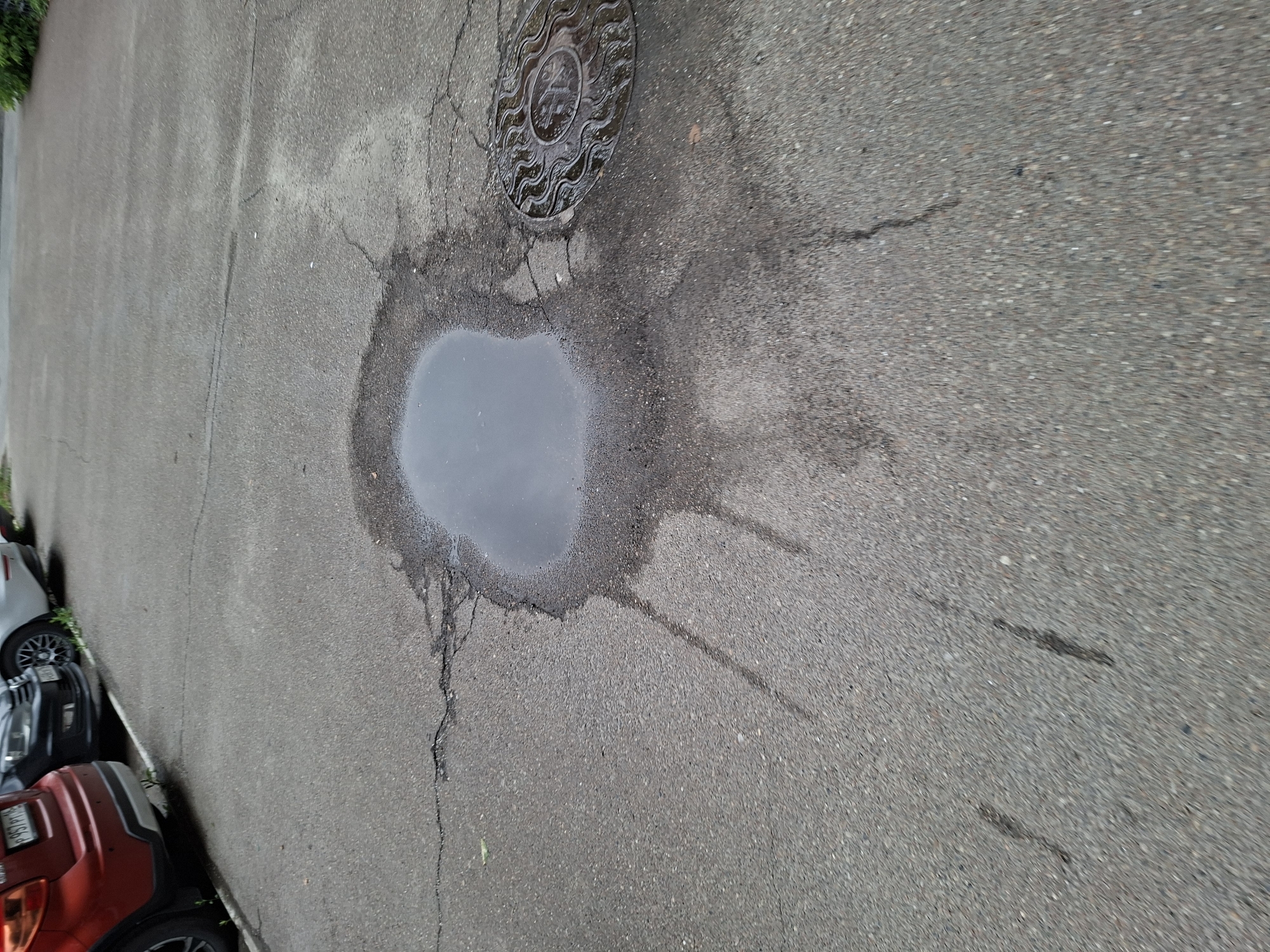 The image shows a puddle of water on a cracked asphalt surface, likely a parking lot or road in Kazan, Russia. The puddle has an irregular shape and appears to be spreading outwards, with streaks of water extending away from the main body. To the right of the puddle, there is a circular manhole cover with a decorative, sun-like pattern. In the background, the rear ends of several cars are visible, including a red SUV on the left. The overall environment suggests an outdoor urban setting.