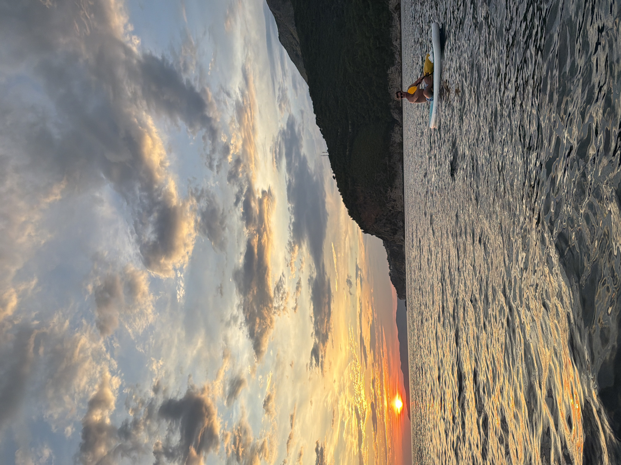 A man is paddleboarding on the calm water near a mountainous coastline during sunset. The sun is low on the horizon, casting a warm, orange glow on the clouds and the water. The sky is filled with dramatic, colorful clouds, ranging from soft grays to vibrant oranges and yellows. The man is seated on his paddleboard, holding a paddle in his hands, and appears relaxed as he enjoys the view. The mountainous landscape behind him is covered in dark green vegetation. The water is calm with gentle ripples reflecting the colors of the sunset. The overall mood of the image is peaceful and serene, capturing the beauty of a sunset at sea. The location is Petrovac na Moru, Montenegro. There is no visible text in the image.