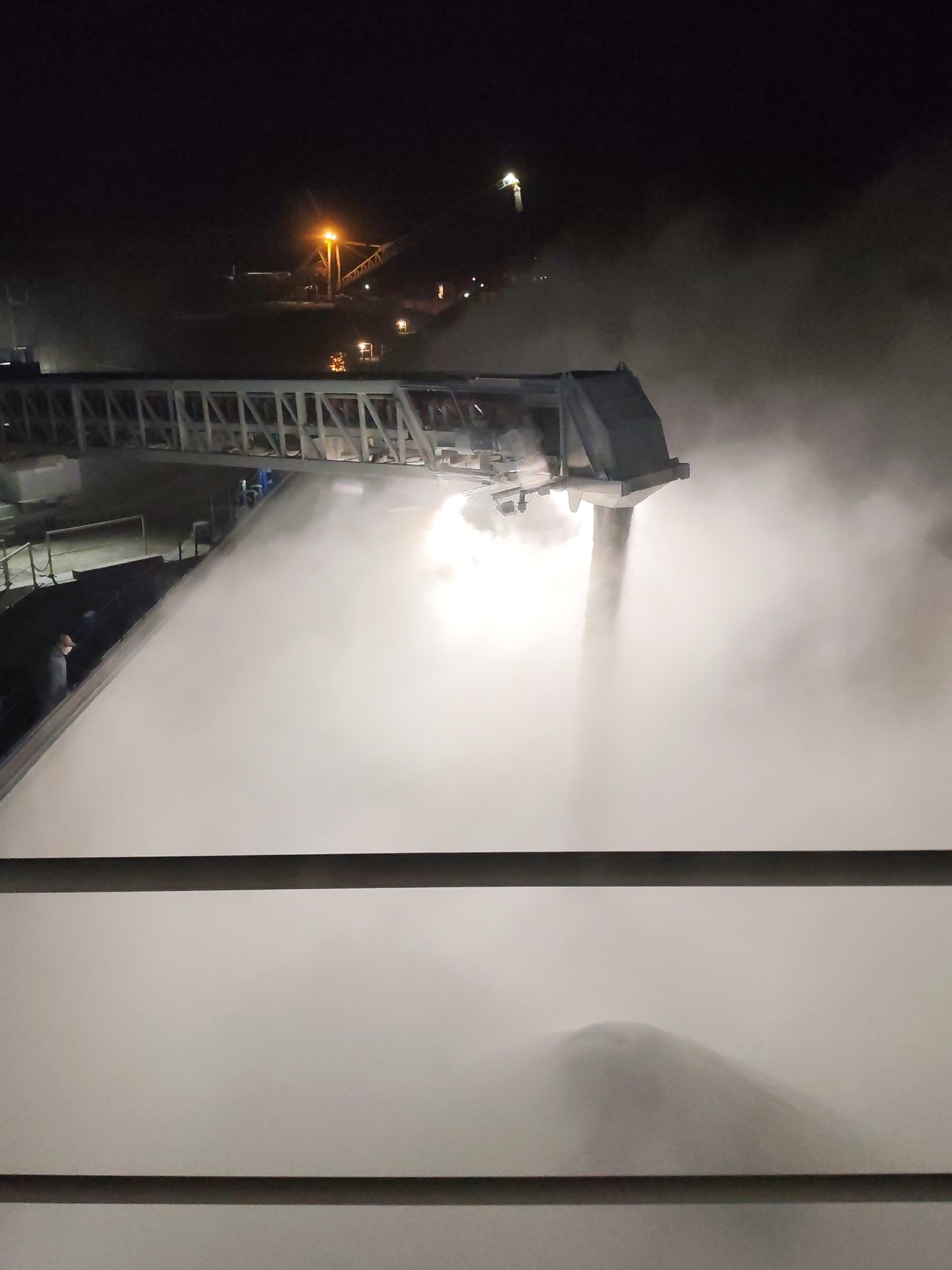 The image depicts an industrial scene at night in Shcheleiki, Russia. An automated conveyor belt system pours a substantial amount of a white, powdery substance (possibly coal or similar material) into a large pile. The process generates a cloud of dust or steam, obscuring much of the view. The structure of the conveyor is visible, with a metal lattice design. In the background, lights from other industrial structures are visible. A single person wearing a mask is partially visible on the left side of the image. The image is taken from an elevated position, looking down at the scene.