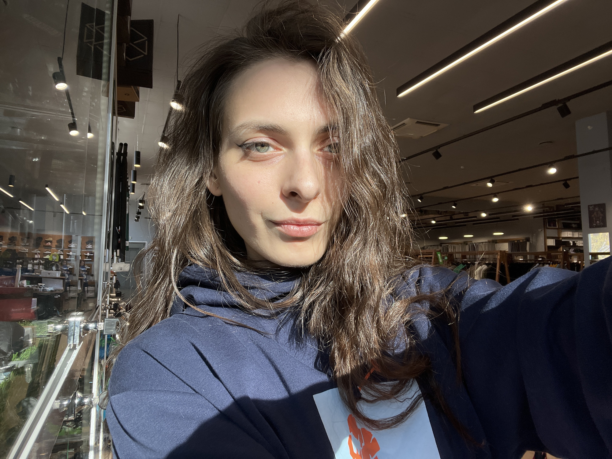 A woman with long, brown, wavy hair wearing a dark blue hoodie takes a selfie in what appears to be a retail store. Her hair is slightly messy. Her skin tone is fair, and she has subtle eyeliner on her eyes. The store features a glass display case to the left, showcasing various objects. Linear LED lighting is visible on the ceiling above. The background suggests an indoor environment with retail displays. Based on available information, the location is the "Триал-Спорт" store in Krasnoyarsk, Russia.