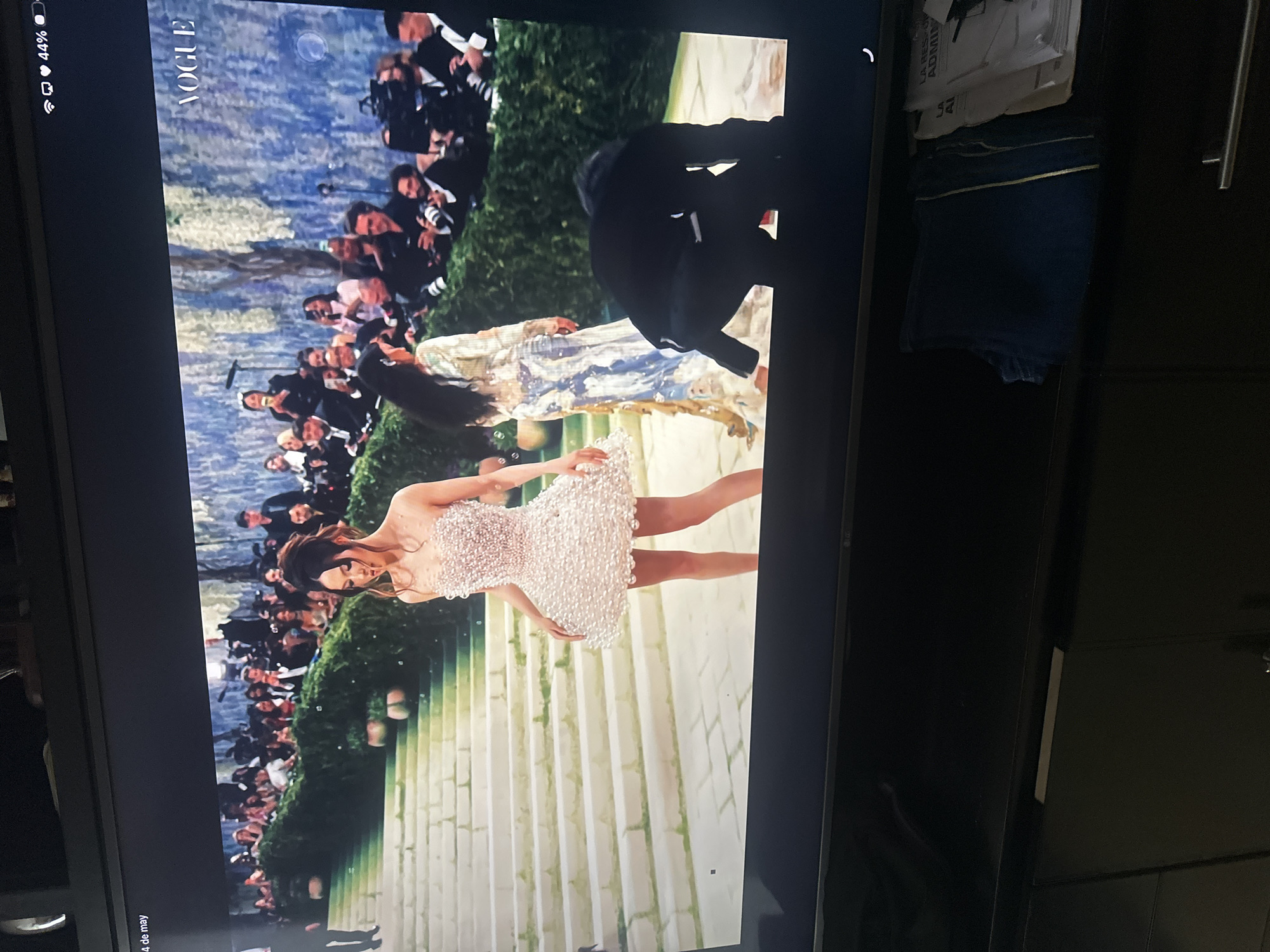 A television screen displays an image of a woman in a white, short dress on a staircase. A crowd of people and photographers are in the background. The image also contains the "Vogue" logo at the top. The location is not Puebla, Mexico. The context appears to be a red carpet or fashion event.