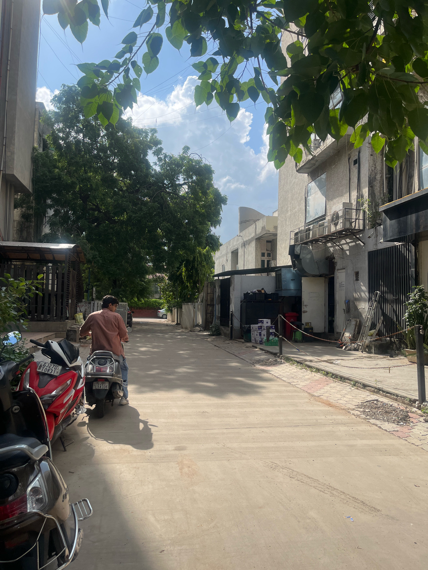 In Navrangpura, India, a street scene unfolds under a bright, partly cloudy sky, with leafy branches of a tree partially obscuring the view. A man wearing a light orange shirt and blue jeans is riding a scooter down a wide, paved street. Parked to his left are three scooters: a red one with license plate GJ27D T0831, a dark-colored one with license plate GJ01 LY4336, and a third dark scooter mostly visible in the foreground. The buildings flanking the street are modern, light-colored structures with visible air conditioning units and large windows. Some buildings appear to be commercial spaces, with metal shutters and open doorways. Debris is scattered along the sidewalk in some areas, and a chain barrier runs along the edge of the paved walkway. The sun casts long shadows on the street, indicating it is likely late morning or early afternoon.