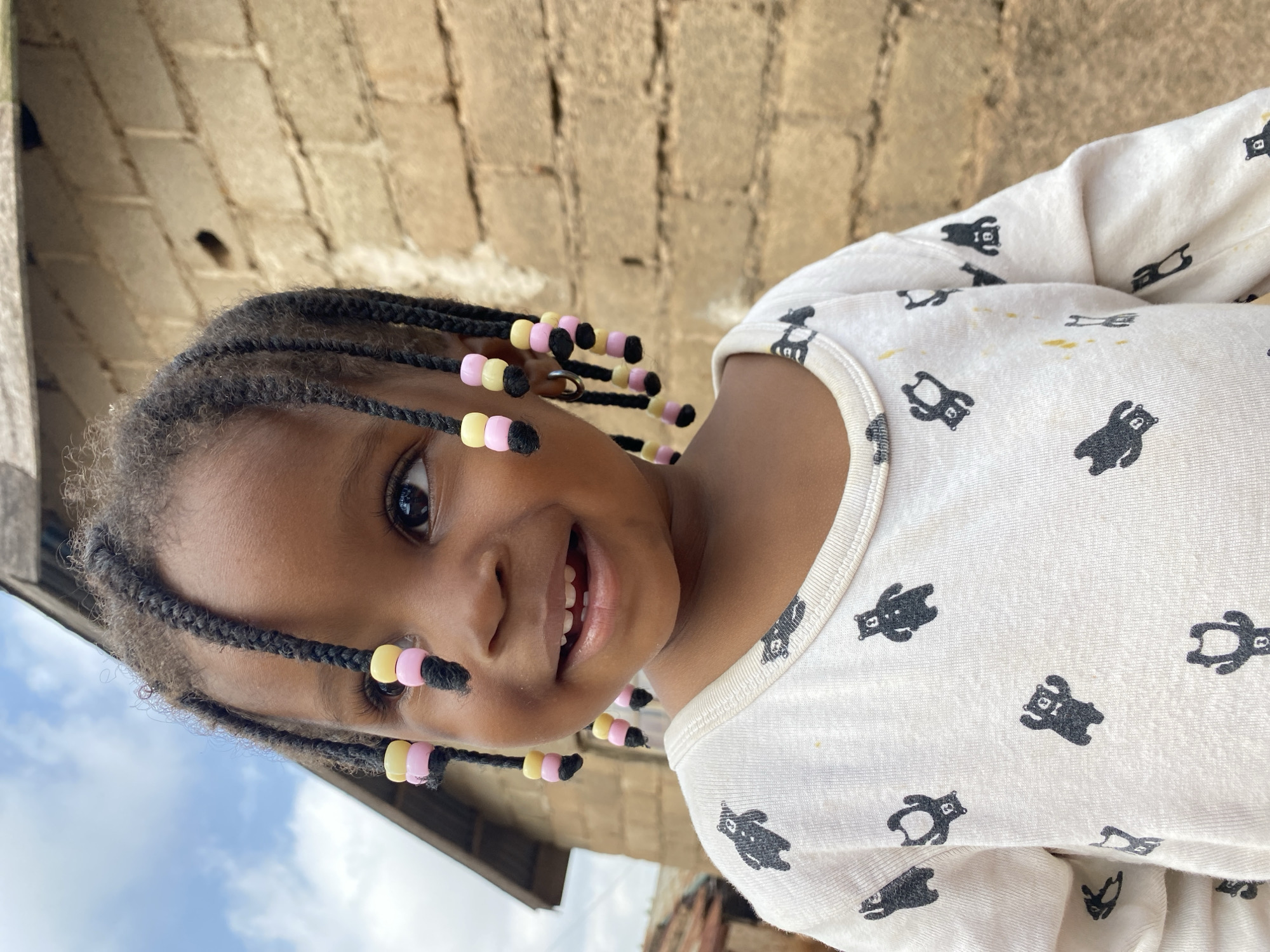 A close-up, eye-level shot shows a young Black child with braided hair adorned with pink and yellow beads. The child wears a white long-sleeved shirt with a pattern of black bear figures. The child is smiling and looking to the left. In the background, a rough, brown brick wall is visible to the right, and a portion of a tin-roofed structure is to the left, under a cloudy sky. The location is the city of Bukuru, Nigeria.