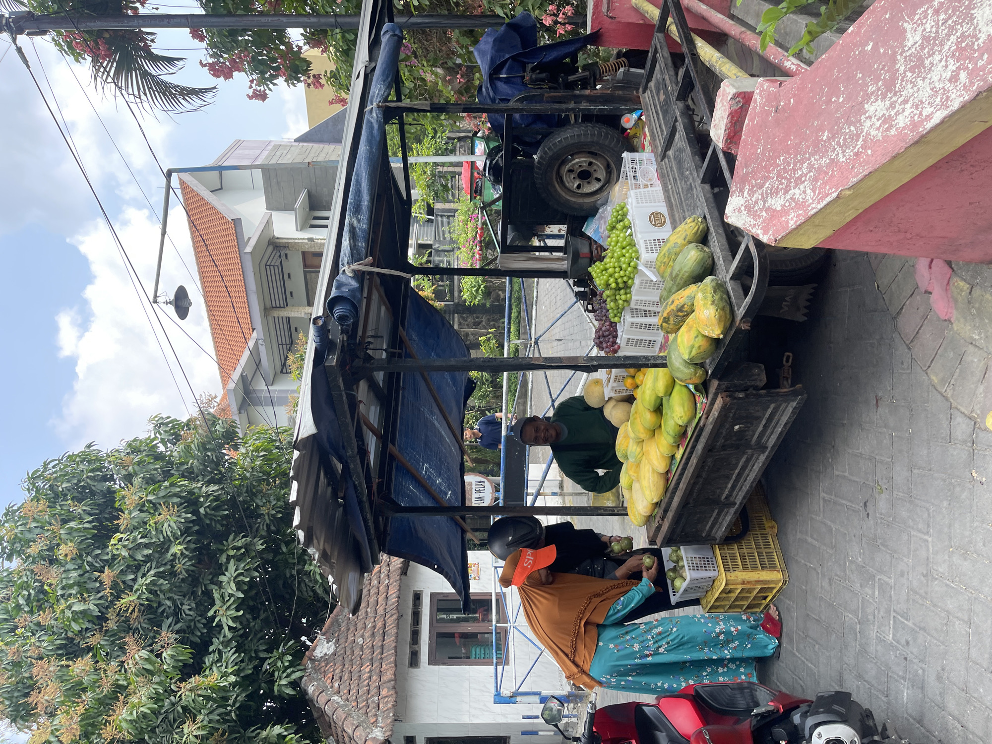 A street fruit stand is set up on a paved road in Larangan, Indonesia, under a blue canopy. The stand is a modified truck with a bed filled with various fruits, including papayas, melons, and grapes, displayed in white crates. A man in a green long-sleeved shirt is standing behind the fruit display, interacting with a woman dressed in a bright orange headscarf and a turquoise dress with a floral pattern. She is holding a basket of green fruits, possibly limes or small guavas, and appears to be engaged in a transaction. Another person in a black helmet stands between them. In the background, a white building with a red tiled roof and a small white gate is visible, suggesting a residential or commercial area. A red scooter is parked to the left of the stand. The scene captures a typical daily activity in a local Indonesian marketplace.