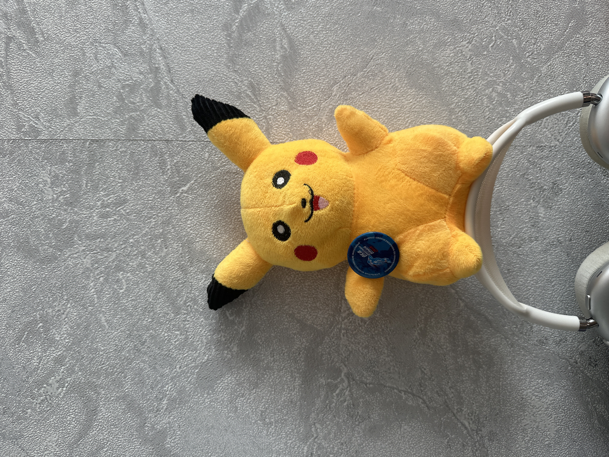 A plush yellow Pikachu toy with black-tipped ears and red cheeks sits atop a white headphone band, which rests on a textured gray wall. The toy is positioned centrally and is facing forward. A small circular blue and white badge is affixed to the Pikachu's chest. The background is a plain, textured gray surface. The scene is devoid of any people.