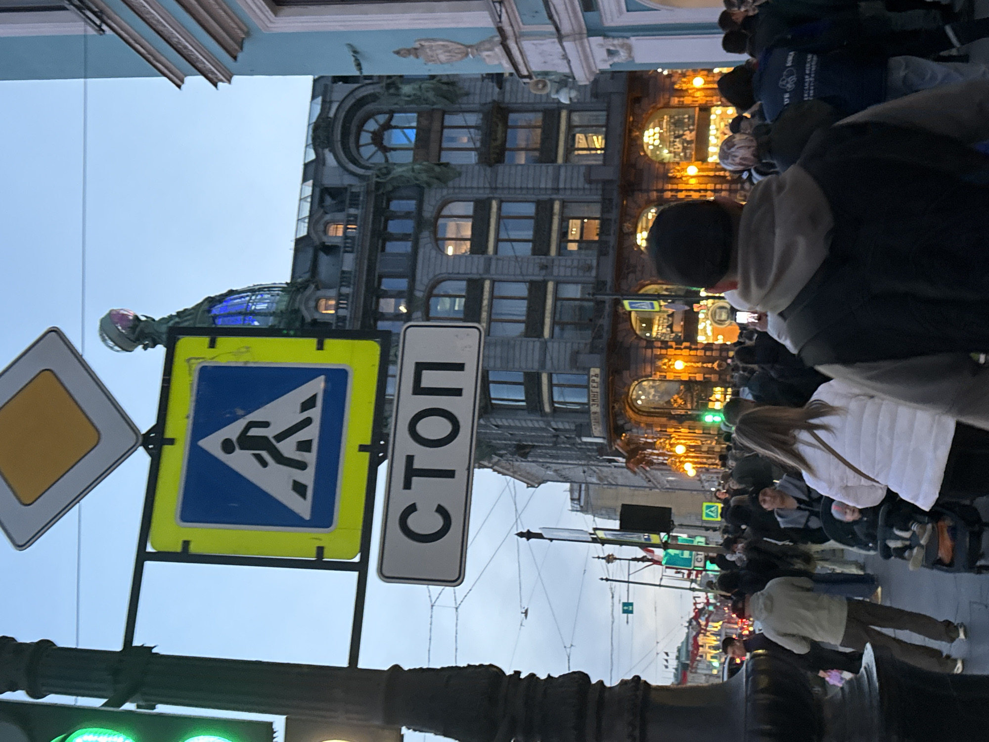 The image depicts a street scene in Saint Petersburg, Russia, in front of the Singer House, also known as the Dom Knigi (House of Books). The building's facade is visible, featuring architectural details such as arched windows, statues, and a glass dome. Street signs are prominently displayed, including pedestrian crossing and stop signs. A crowd of people is present on the sidewalk, walking in both directions, and some are likely tourists. The light suggests that the photograph was taken during daytime, under a cloudy sky.