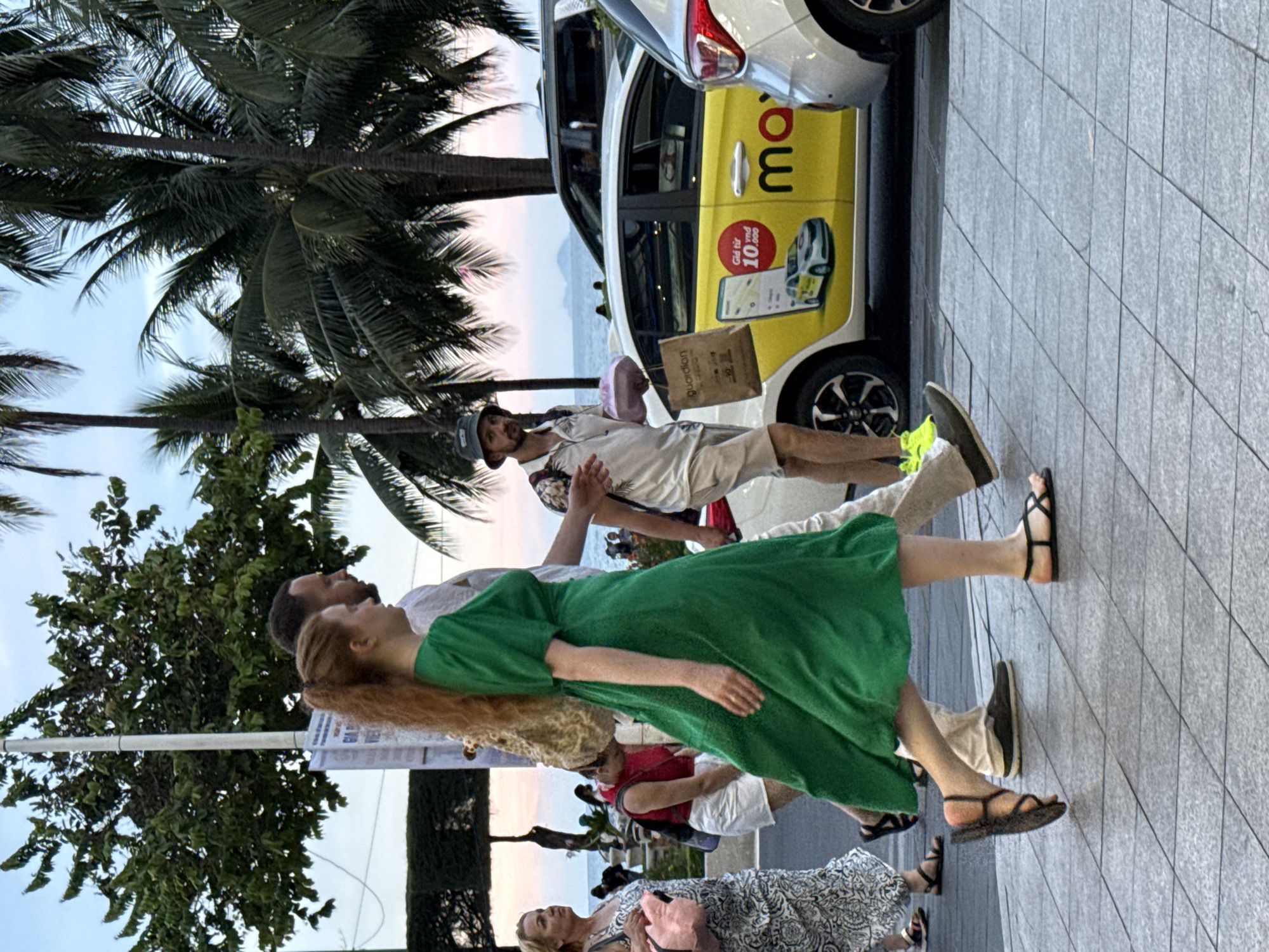 The image shows a street scene in Nha Trang Ward, Vietnam, with several people walking along a tiled sidewalk. A woman in a green dress and a man in a light-colored shirt and shorts are walking towards the right, with the woman in the foreground. To the left, another woman in a patterned dress is walking, carrying a pink bag. Further back, a man wearing a hat and carrying bags is walking, with a yellow taxi visible behind him. Palm trees line the background, and the ocean is visible in the distance. The sky is a pale blue with hints of pink, suggesting it is either early morning or late evening.