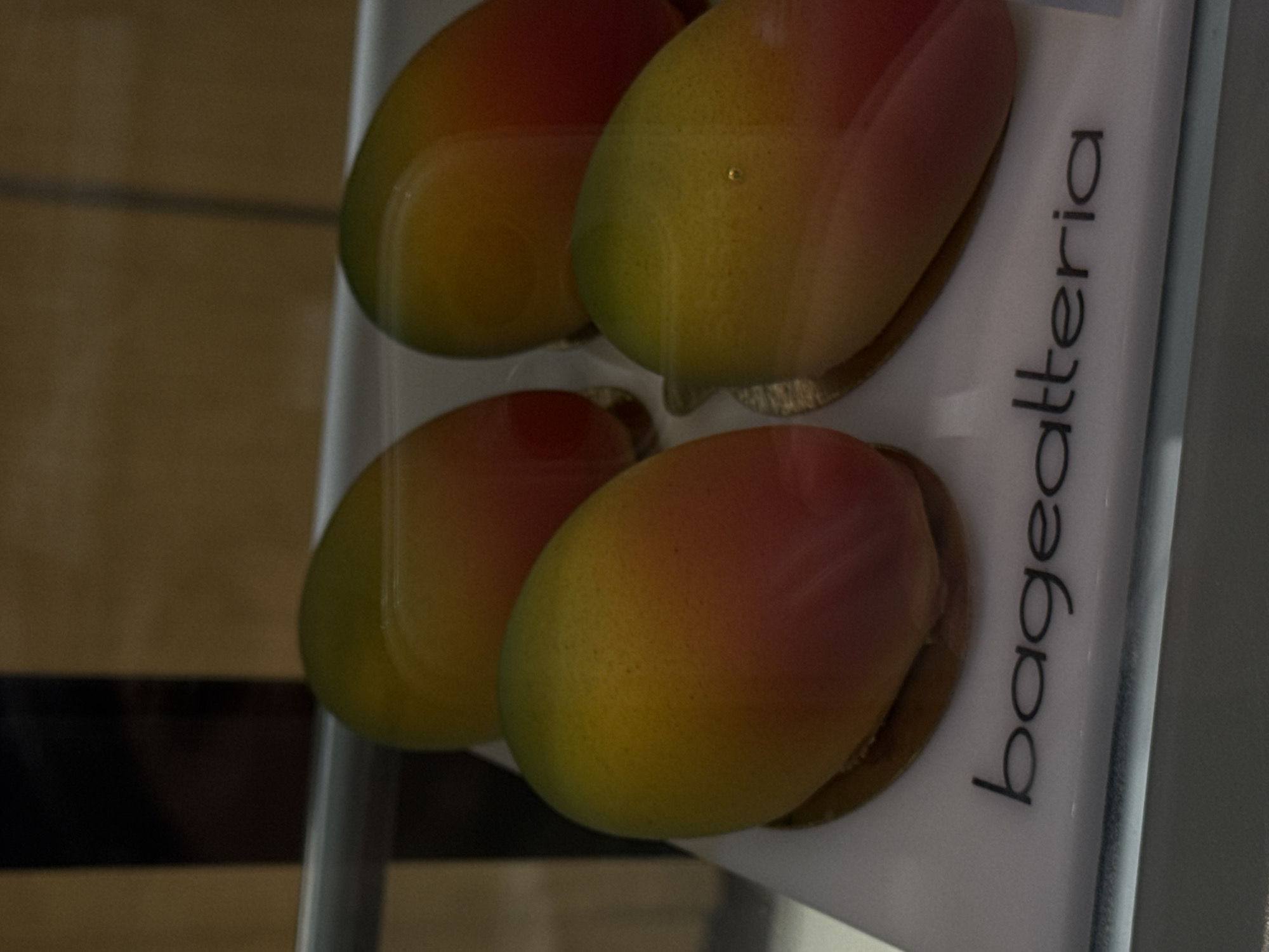 A close-up shot shows four mango-shaped objects encased behind a clear plastic display case. The objects are arranged in two rows of two. The mangoes on the left are located near a dark brown wooden surface. The case is attached to a white display with black writing, reading "bageatteria". The image was taken in a location within Moscow, Russia.