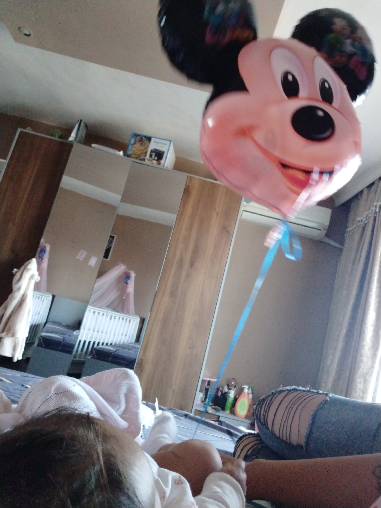 The image shows a baby lying on a bed, presumably in a bedroom. A large Mickey Mouse balloon floats in the air, held by a blue ribbon. A person wearing ripped jeans is visible in the lower right corner of the image. In the background, there is a wardrobe with mirrored doors that reflects a baby's crib covered with a pink net, suggesting a nursery setting. Some toiletries are seen on a shelf in the background, and curtains hang by the window. The location is the city of Unknown, Unknown.