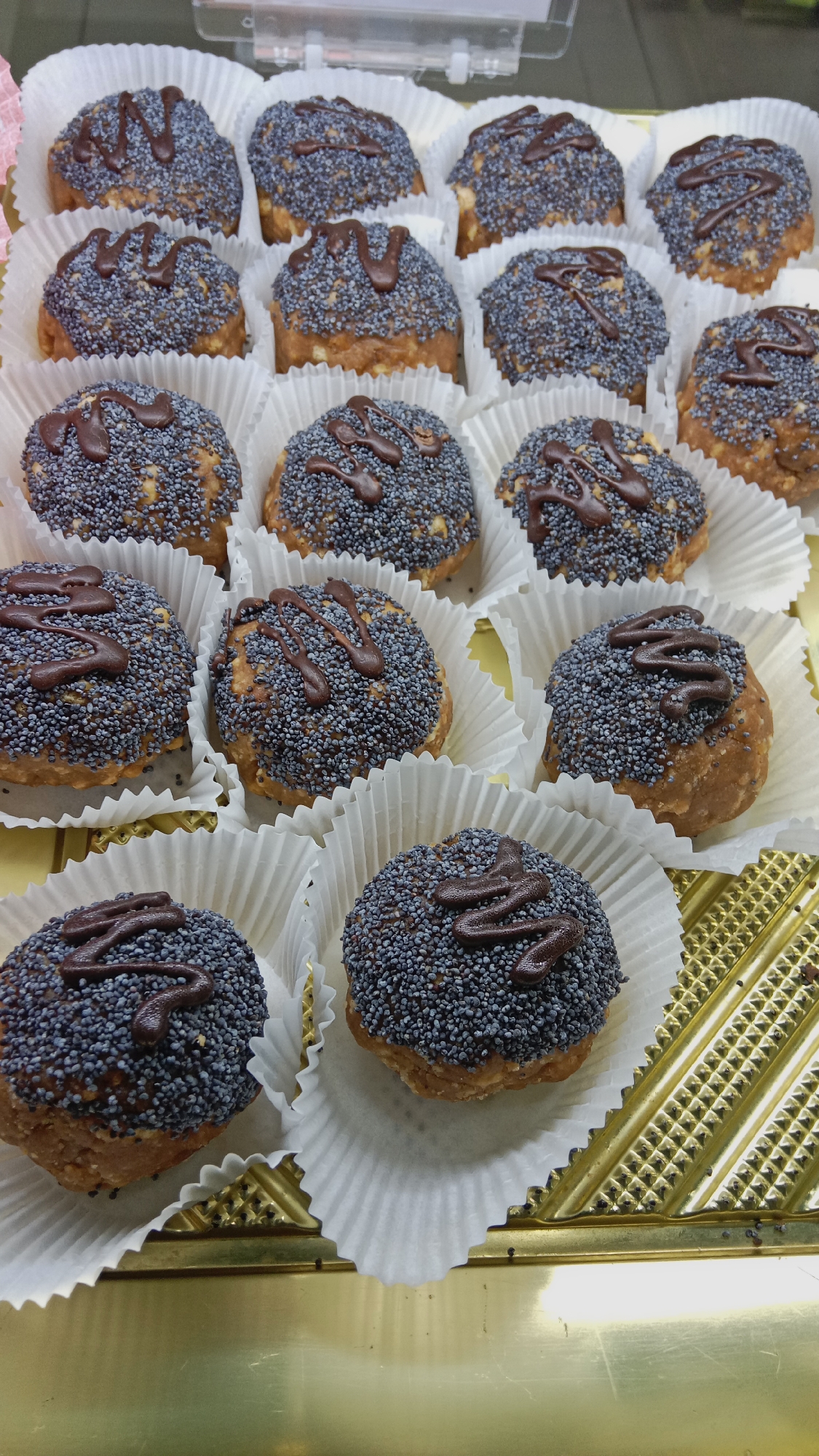 A close-up, overhead shot displays a tray of approximately 15 poppy seed pastries, each presented in a white paper cupcake liner. The pastries are round and topped with a dense layer of poppy seeds and a drizzle of dark chocolate. They appear to be arranged in a grid formation within a golden-colored tray, suggesting they are for sale in a bakery or confectionery. The background is slightly blurred, showing hints of a retail environment. The pastries are the primary focus, with their textures and arrangement clearly visible. The lighting is even, highlighting the details of the baked goods.