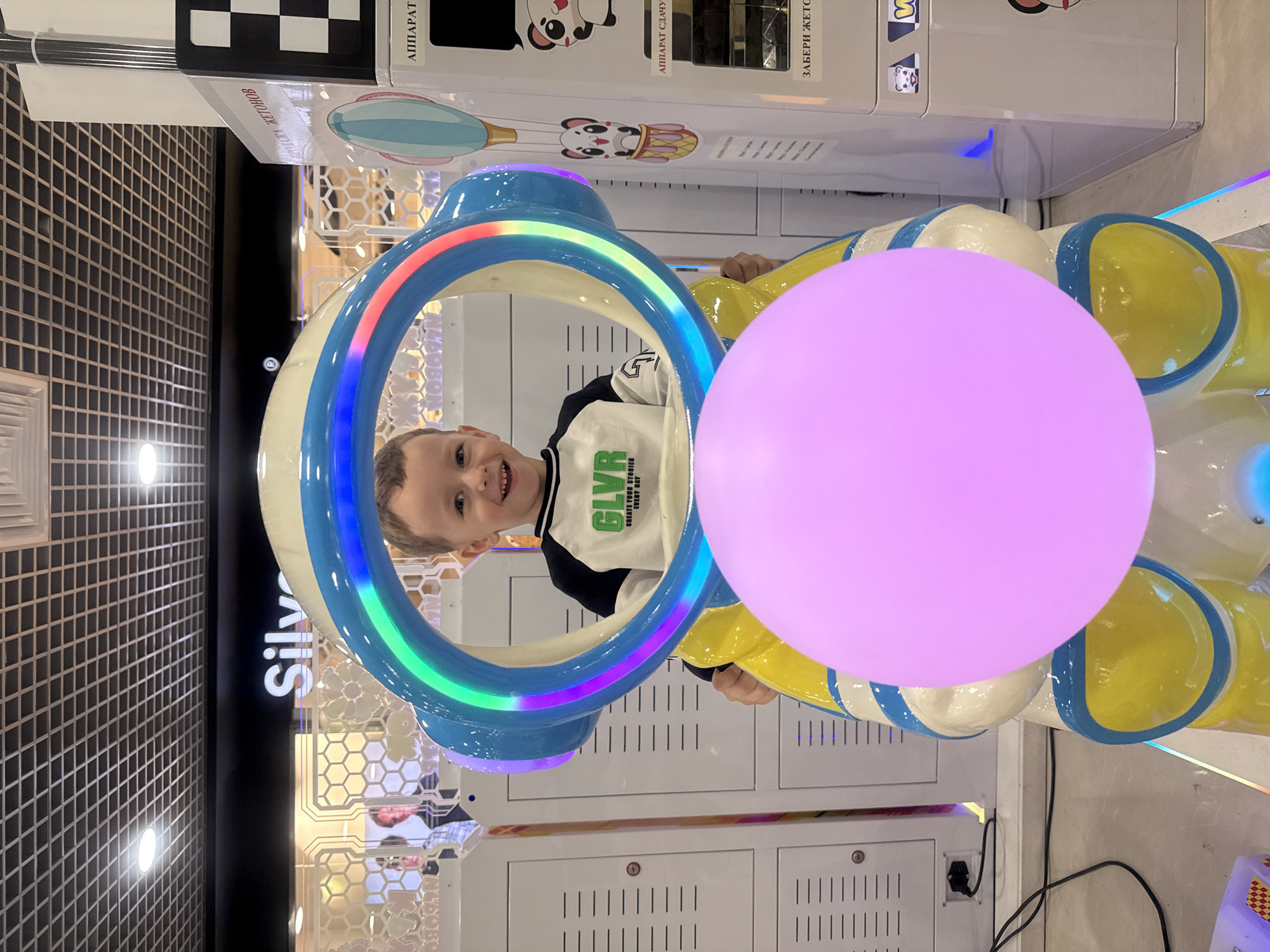 A young male child, approximately 4-6 years old, is seated on a brightly lit amusement ride within an indoor entertainment center in Vladivostok, Russia. The child, with light brown hair and a white-and-dark-blue long-sleeved shirt bearing green text "GLVR CREATE YOUR STORIES EVERY DAY," smiles broadly directly at the camera. He is positioned behind a large, arch-shaped element of the ride, which is illuminated with multicolored LED lights displaying a rainbow spectrum. A prominent, glowing spherical pink object is situated directly in front of the child, obscuring the lower portion of his body and the base of the ride.
The ride itself is predominantly white, light blue, and yellow. The background reveals a typical arcade environment with various white game machines or vending units. On the left, a partial sign reads "Sily" against a dark band, above a patterned glass partition reflecting two indistinct figures. On the right, white machines feature Cyrillic text including "ПРОДАЖА ЖЕТОНОВ" (Token Sale) and "АППАРАТ СДАЧИ" (Change Machine), alongside cartoon panda graphics and a hot air balloon motif. The ceiling features a dark grid pattern with recessed circular lights. The floor is light-colored, showing reflections from the ride's lights, with a visible black power cord on the left.