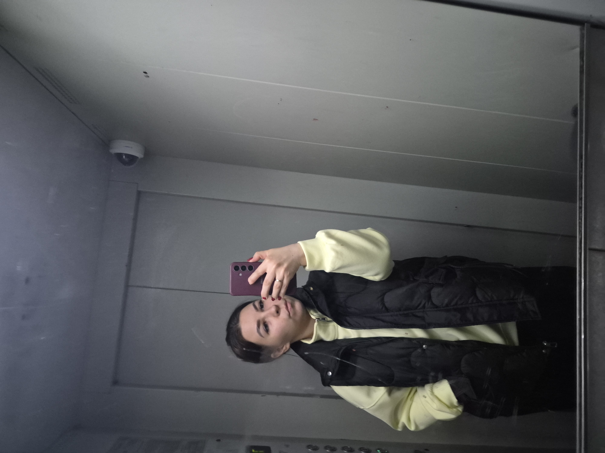 A person stands in what appears to be an elevator, taking a selfie using a purple smartphone. The individual is wearing a light yellow hoodie and a black vest. A security camera is mounted in the top left corner of the elevator. The elevator's interior walls are gray and metallic. The location is potentially in Tobolsk, Russia.