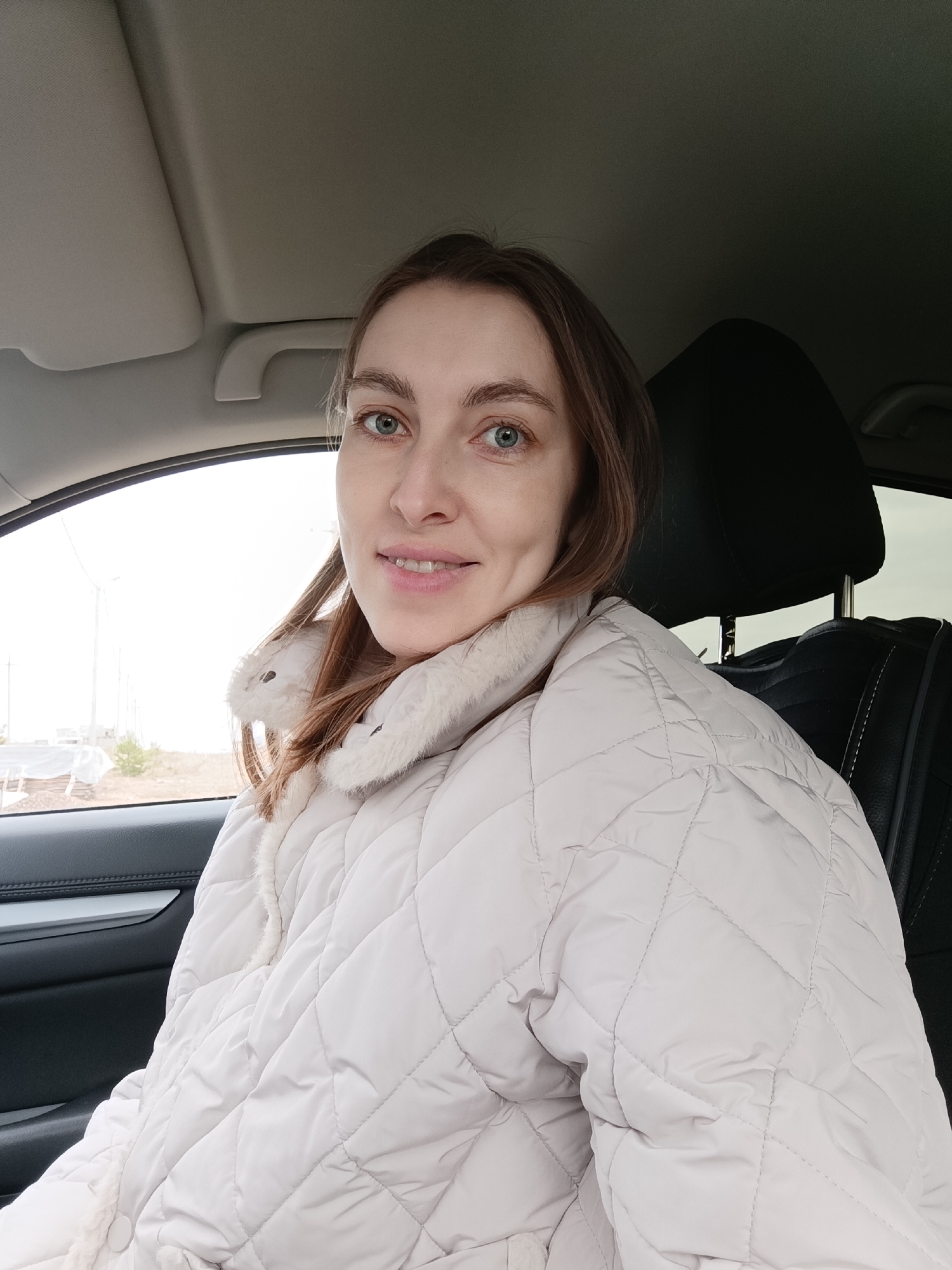 The image depicts a woman in a car. She has fair skin, brown hair, and blue eyes. She is wearing a light-colored, quilted jacket with a fur-lined collar. The interior of the car is visible, including the black leather seats and the car's interior trim. Outside the window, a flat landscape is visible. Based on the background details, it is impossible to confirm the location in Chaikovsky, Russia.