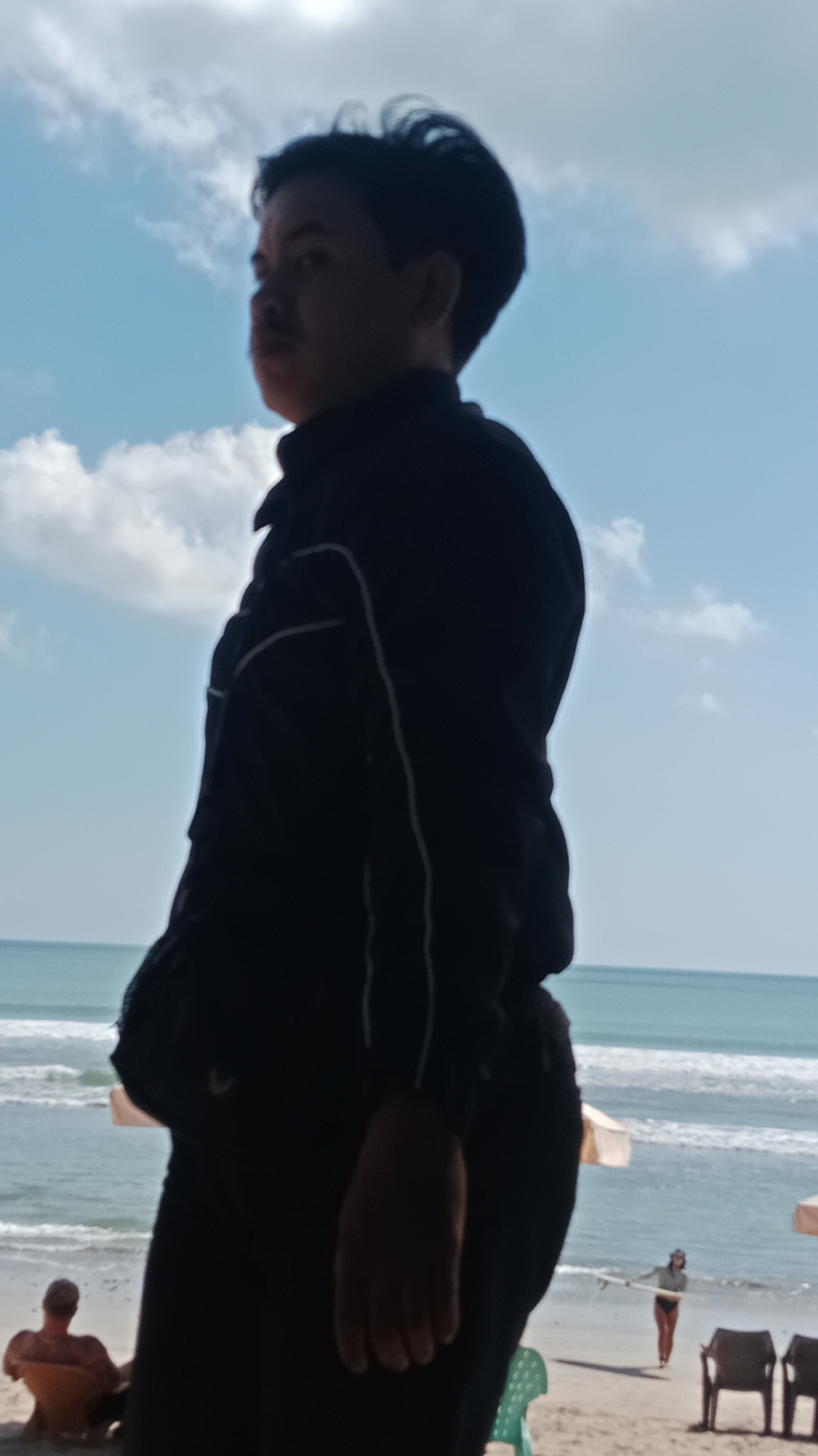 The image shows a silhouette of a person in the foreground, facing away from the camera and looking out at the ocean. The person is wearing dark clothing. In the background, there is a beach with several other people visible. One person is sitting on a chair facing the ocean, and another person is walking with a surfboard. There are also umbrellas and chairs on the beach. The ocean is calm with small waves. The sky is blue with some white clouds. The scene appears to be a beach in Kuta, Indonesia, given the general appearance of the beach and the ocean.