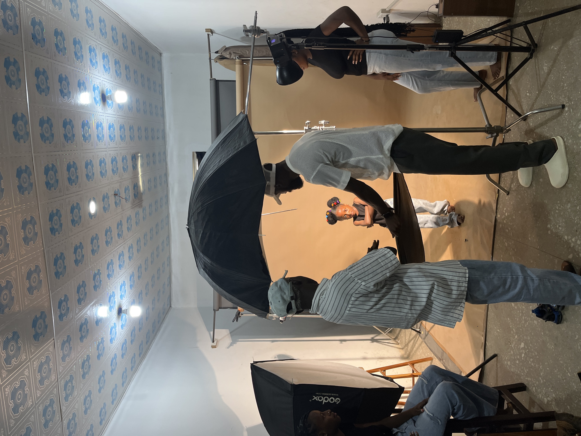 This is a forensic, matter-of-fact description of a photography studio scene in Gura Top, Nigeria. The image depicts a professional studio setting with a backdrop of a light brown seamless paper roll, suspended by a lighting stand and a horizontal bar. The ceiling is decorated with a patterned tile design.
In the foreground, four individuals are present, engaged in what appears to be a photoshoot. To the left, a woman is seated on a dark wooden stool, wearing a black t-shirt and light blue distressed jeans. Next to her, facing away from the camera, is an older man in a striped shirt and light blue pants. He is gesturing with his hands towards a young girl.
The central subject of the photograph is a young girl, positioned between the older man and another adult. She is wearing a black dress and has her hair styled in two buns with colorful accessories. She stands with her arms crossed, looking towards the camera.
To the right of the girl, an adult male, facing away from the camera, is wearing a white mesh shirt and dark pants. He stands near the girl, potentially directing her pose. Further to the right, an adult female stands with her hands on her hips, wearing a black t-shirt and light blue jeans. She is looking towards the girl.
Various pieces of photography equipment are visible, including a large black umbrella positioned above the girl, a softbox on the left, and a studio light on a tripod to the right. The floor is a light gray concrete with dark speckles. The overall lighting suggests a controlled studio environment.