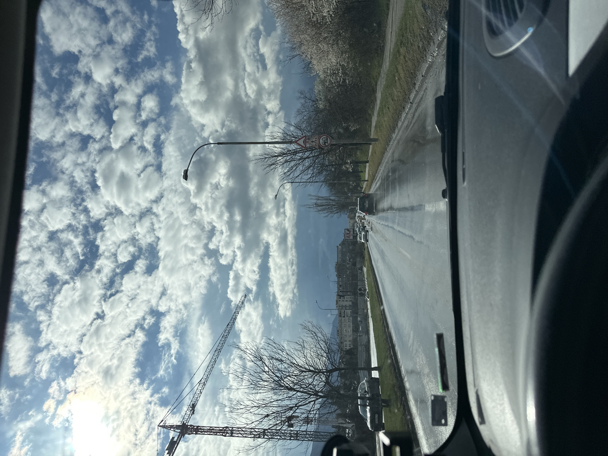 The image, taken from inside a vehicle, shows a road in Sofia, Bulgaria, on a wet day. Rain streaks are visible on the windshield. The road has multiple lanes with moving traffic. Alongside the road is a grassy area and trees with bare branches. Further in the background, buildings and a crane can be seen. The sky is blue with scattered white clouds. A speed limit sign is visible next to a tree.