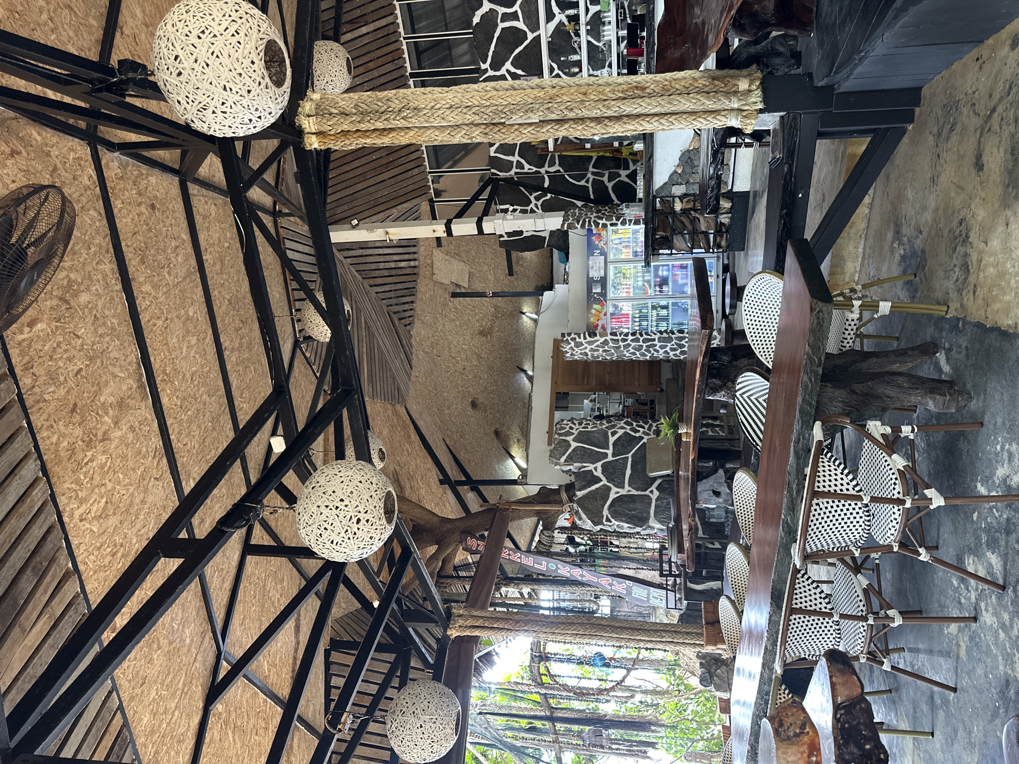 This is an interior shot of a rustic restaurant in Ko Pha Ngan, Thailand, characterized by its open-air design and natural materials. The ceiling features exposed wooden planks and a dark metal framework, supporting several large, white, woven spherical light fixtures. A ceiling fan is visible in the upper left. The main seating area consists of several large, live-edge wooden tables, with chairs upholstered in a black and white checkered pattern and wicker. One table on the left has a rough-hewn wooden base. In the background, a bar area is visible with refrigerators stocked with beverages, and shelving units displaying bottles. The walls behind the bar are decorated with a black and white stone or tile pattern. Large, thick ropes are used as decorative elements and structural supports, adding to the tropical, bohemian aesthetic. Natural light filters in from the sides, and lush green foliage is visible through openings in the structure. The floor is a rough concrete or stone surface. There are no people visible in the image.