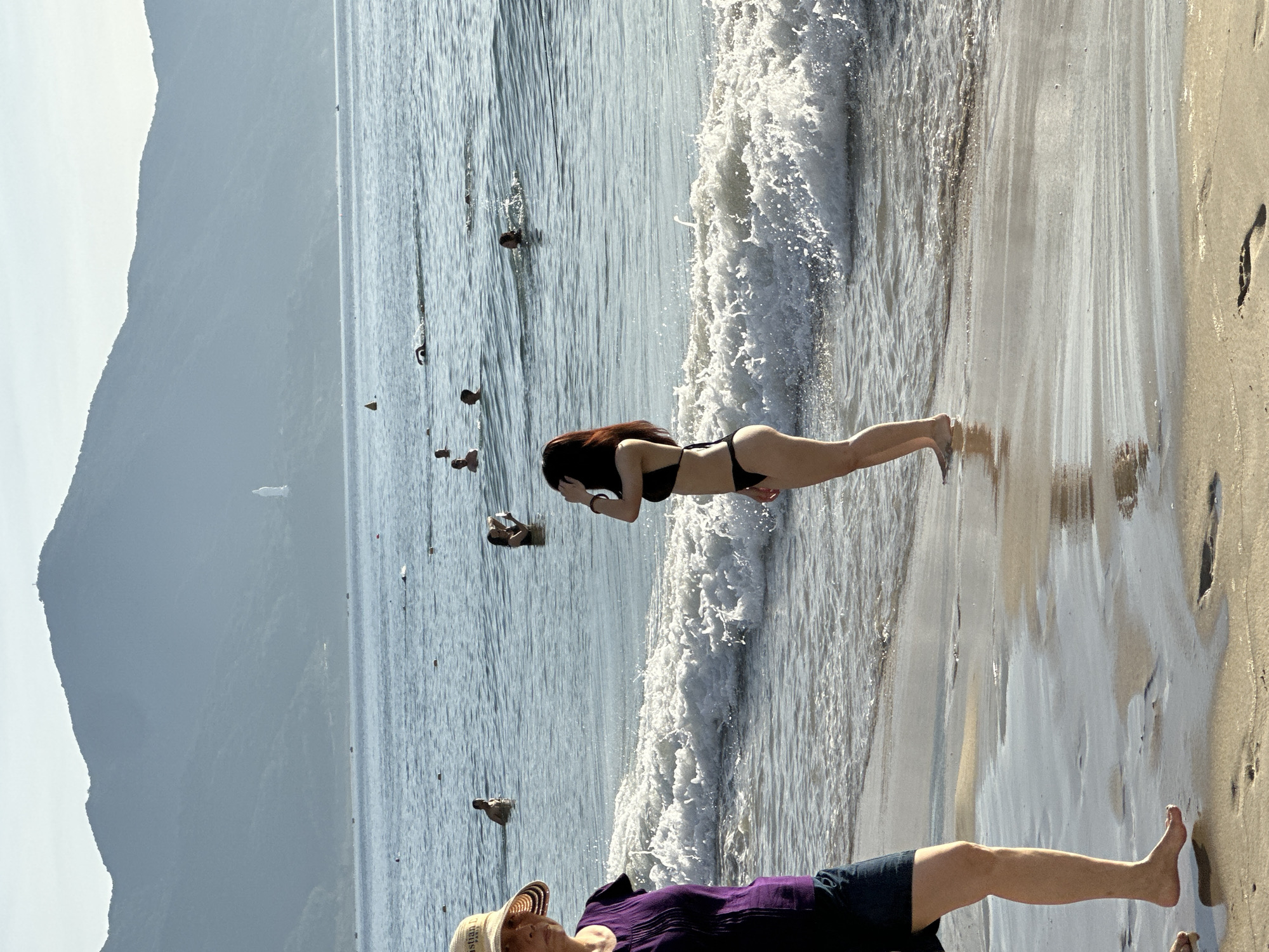 The image captures a coastal scene at a beach in Đà Nẵng, Vietnam, likely Mỹ Khê Beach given its popularity and typical mountainous backdrop. The sun is setting, casting a warm, soft light on the water and sand. In the foreground, a woman wearing a purple shirt and dark shorts stands on the beach, facing away from the camera and towards the ocean. To her right, another woman in a black bikini is partially visible, standing in the shallow water.
The ocean is calm with gentle waves breaking on the shore, creating a foamy white line. In the middle ground, several people are visible swimming and enjoying the water. The background is dominated by a large, verdant mountain range that stretches across the horizon, its peaks softened by the hazy light. A faint white sail of a boat can be seen in the distance on the water. The overall atmosphere is peaceful and relaxed, typical of a late afternoon at a popular Vietnamese beach.