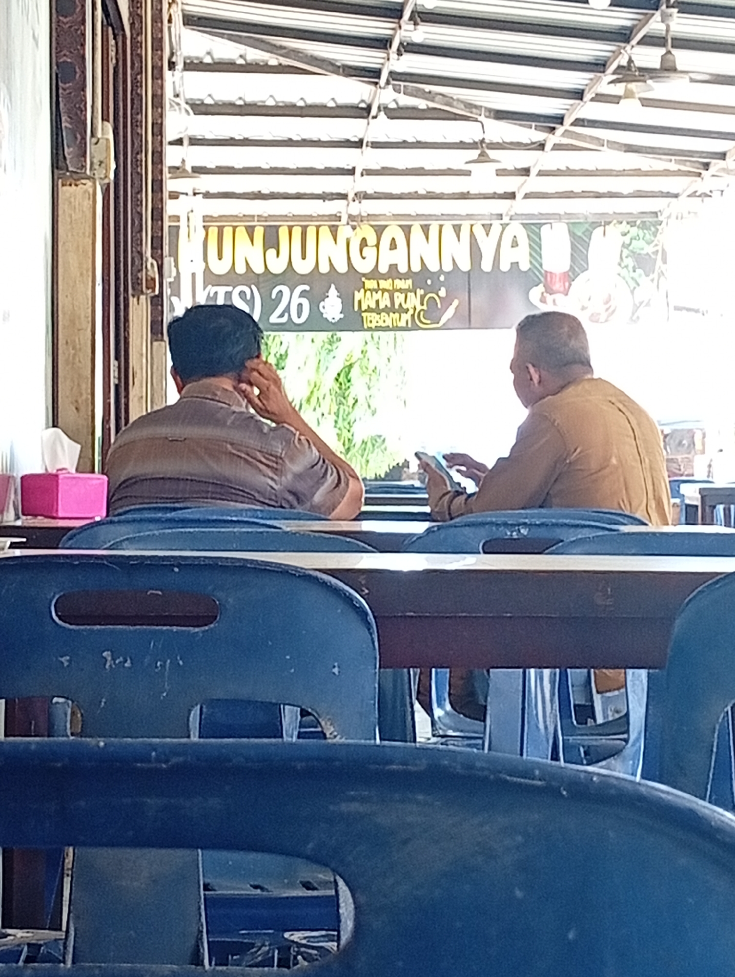 The image shows two men sitting at a table in what appears to be an outdoor dining area. The man on the left is wearing a brown and gray striped shirt and has his hand to his ear. The man on the right is wearing a brown shirt and is looking at his phone. In the background, a sign reads "KUNJUNGANNYA" with other text below it. There are blue chairs and a wooden table in the foreground. A pink tissue box is visible on the table to the left. The location is identified as Lhokseumawe, Indonesia.