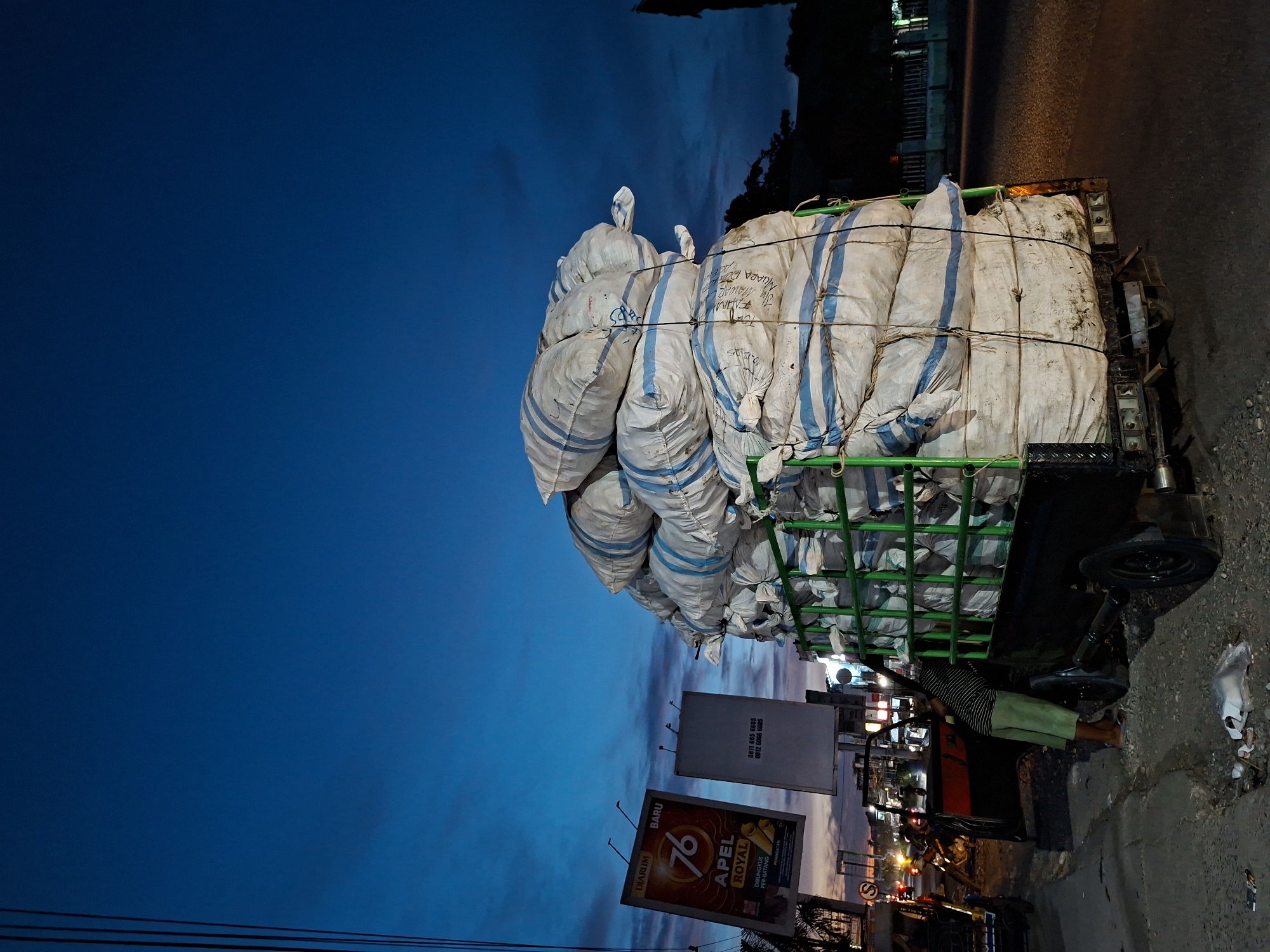 The image captures a scene in Lhokseumawe, Indonesia, at twilight, with a large, precarious stack of white sacks secured with blue-striped bands, piled high on the back of a green-framed truck. The sacks fill the upper portion of the image, contrasting with the deep blue of the evening sky. Below, the truck is parked along a paved street, where a person is positioned near the rear tire. Adjacent to the truck, an advertising billboard featuring "DIARLIM BARU 76 APEL ROYAL" is visible, with contact numbers displayed. Streetlights and other city illuminations punctuate the background, indicative of an urban environment. Electrical wires stretch across the lower-left corner of the frame. The roadway shows signs of wear and debris.