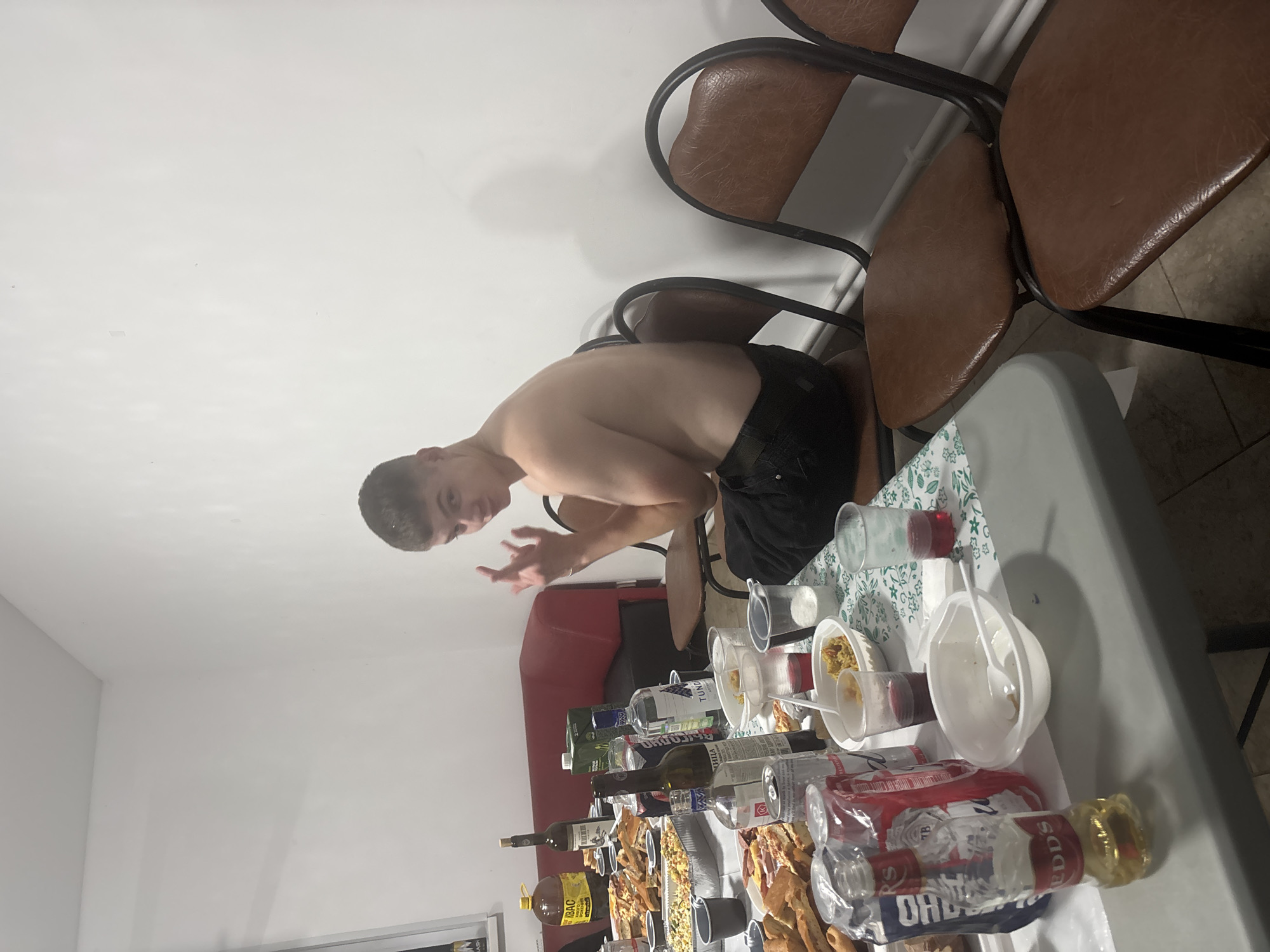 In a dimly lit room, a young man with short dark hair, shirtless and wearing black shorts, is the central subject of the image. He is captured mid-motion, leaning forward with his right arm extended and his hand open, as if speaking or gesturing. His expression is animated, suggesting engagement in a conversation or activity.
The scene unfolds around a table laden with food and drinks, indicating a gathering or party. Several bottles of alcohol are visible, including what appears to be a bottle of vodka with a label featuring blue and white text, and another bottle with yellow liquid and a red label that reads "IBAC". There are also various containers, some filled with food like snacks or appetizers, and disposable cups. A white plastic bowl with a spoon suggests recent use.
The setting appears to be an indoor space, possibly a dining or common area, with a plain white wall in the background and a portion of a red sofa visible. Several chairs with brown leather seats and dark metal frames are positioned around the table, hinting at a comfortable but casual environment. The overall atmosphere seems lively and informal, typical of a social event.
Although no specific time of day is explicitly indicated, the lighting suggests it could be evening or nighttime. The image is a snapshot of a moment, capturing the energy of the young man and the convivial atmosphere of the gathering. No overt weather cues are present, as it is an indoor scene.