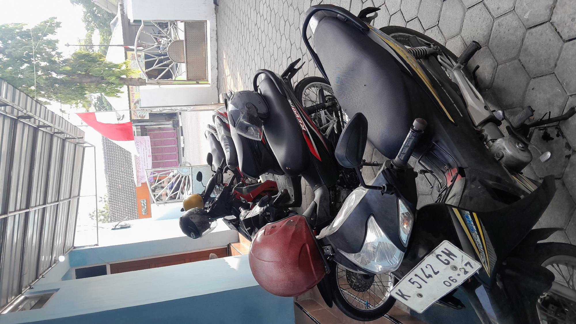 This is a medium shot, eye-level, outdoor photograph of several parked motorcycles in Sambong, Indonesia. The motorcycles are lined up parallel to a building with a light blue wall and a gray metal roof overhang. The ground is paved with hexagonal gray tiles. In the background, a red and white Indonesian flag flies from a flagpole. There are also buildings visible, one with a tiled roof and another with pink doors. Some of the motorcycles have helmets on them. One motorcycle in the foreground has a license plate that reads "K 5142 GH 06-27". The overall scene suggests a street or an area in front of a business or residence.