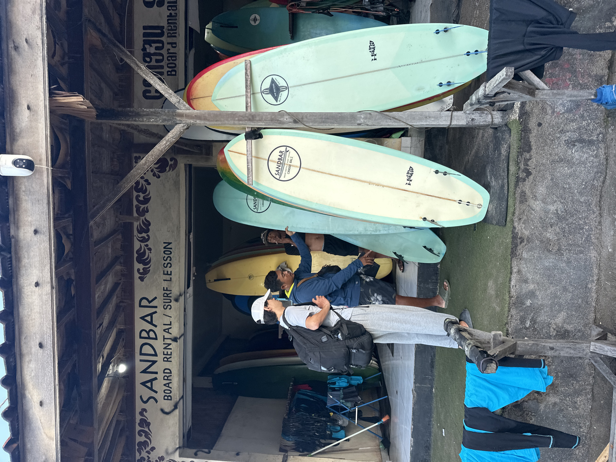 In North Kuta, Indonesia, a surf shop named SANDBAR, which offers board rentals and surf lessons, is depicted. The shop front features a rustic wooden overhang with visible beams and a thatched roof. A white security camera is mounted on the overhang. Below the overhang, a sign with "SANDBAR" in large letters is visible, along with smaller text indicating "BOARD RENTAL / SURF LESSON".
Several people are present. Two young men are standing near the entrance, looking at surfboards. One, wearing a white cap, grey t-shirt, and grey sweatpants, has a large black backpack. The other, in a blue hoodie and patterned shorts, appears to be gesturing towards the surfboards. Another man is standing further back, partially visible, holding a cigarette. A few other surfboards are leaning against the shop.
On the left side of the image, a rack of black and blue wetsuits or surf attire hangs on a wooden frame. The ground in front of the shop is a mixture of tiled flooring and green artificial turf. Various surfboards, in different colors including light blue, yellow, and mint green, are lined up, some with logos like "SANDBAR" and "Hana". The overall scene suggests a casual, outdoor environment typical of a beachside town.