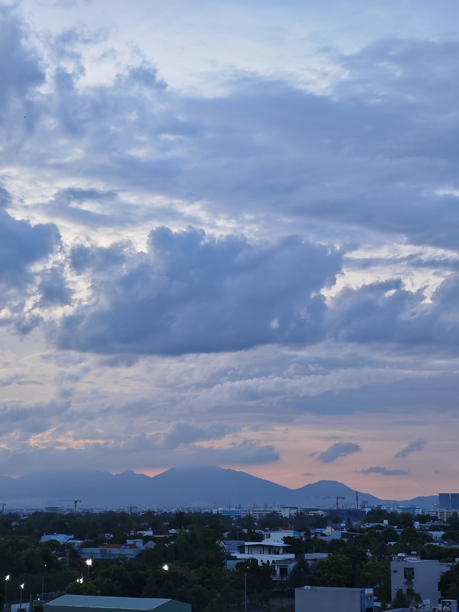This is an eye-level, medium shot of a city skyline at dusk. The sky is filled with dramatic, swirling clouds in shades of gray, white, and hints of pink from the setting sun. In the foreground, a dense canopy of dark green trees obscures most of the residential buildings, with only the tops of some houses and structures visible. Beyond the trees, the city unfolds with a mix of low-rise buildings, industrial cranes, and taller, modern structures. In the distance, a range of majestic, blue-hued mountains forms a serene backdrop against the colorful sky. The overall atmosphere is peaceful and serene, with the fading light casting a soft glow over the urban landscape. This scene is likely the city of Đà Nẵng, Vietnam, given the characteristic mountain range and urban development.