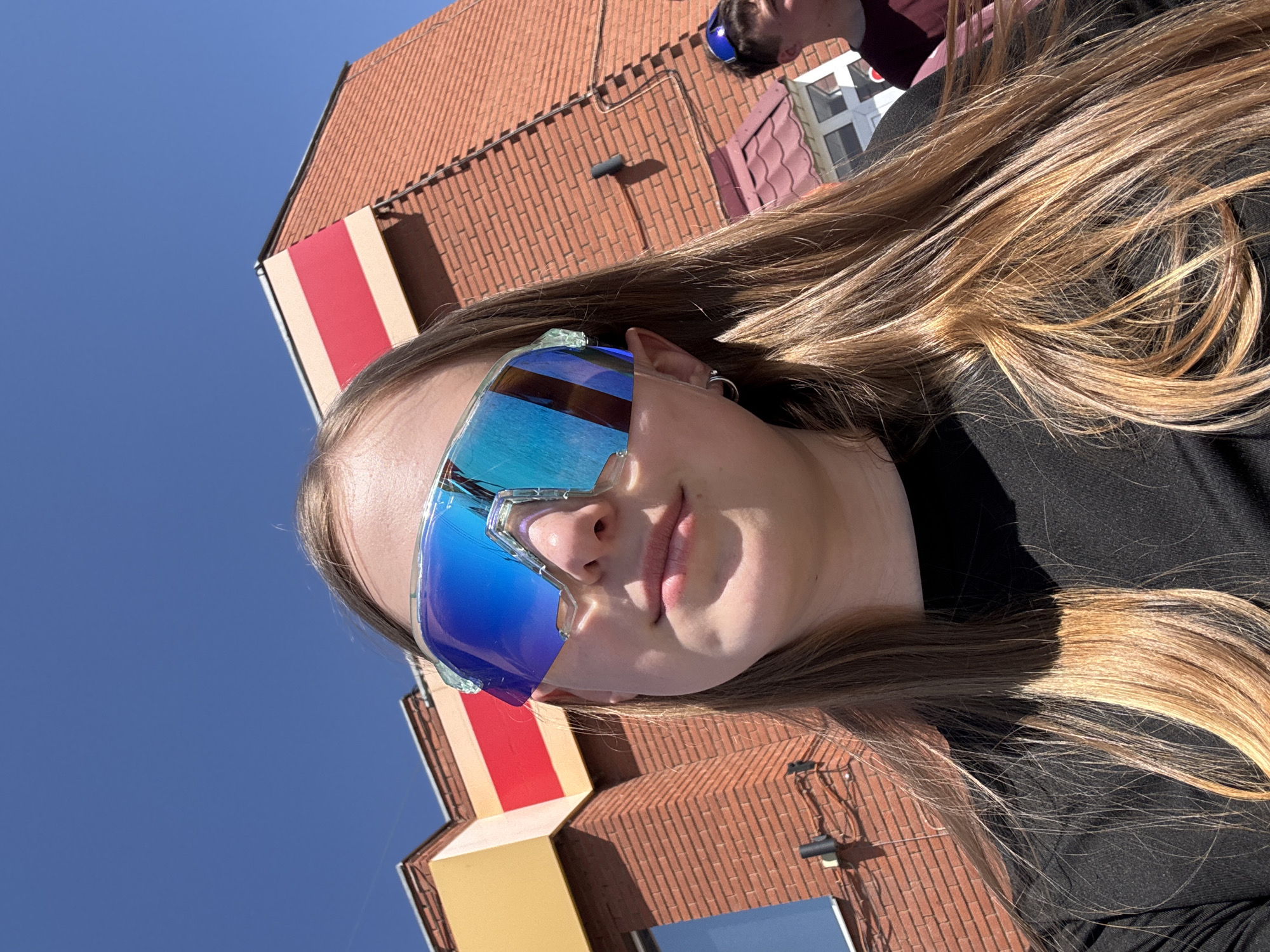 A medium shot captures a woman with long blonde hair wearing oversized blue mirrored sunglasses. She is outdoors, angled upward toward a clear blue sky. Behind her, buildings with brick facades and red and white striped elements are visible. Another person wearing sunglasses is partially visible in the background, out of focus. The location is possibly in Grishanki, Russia.