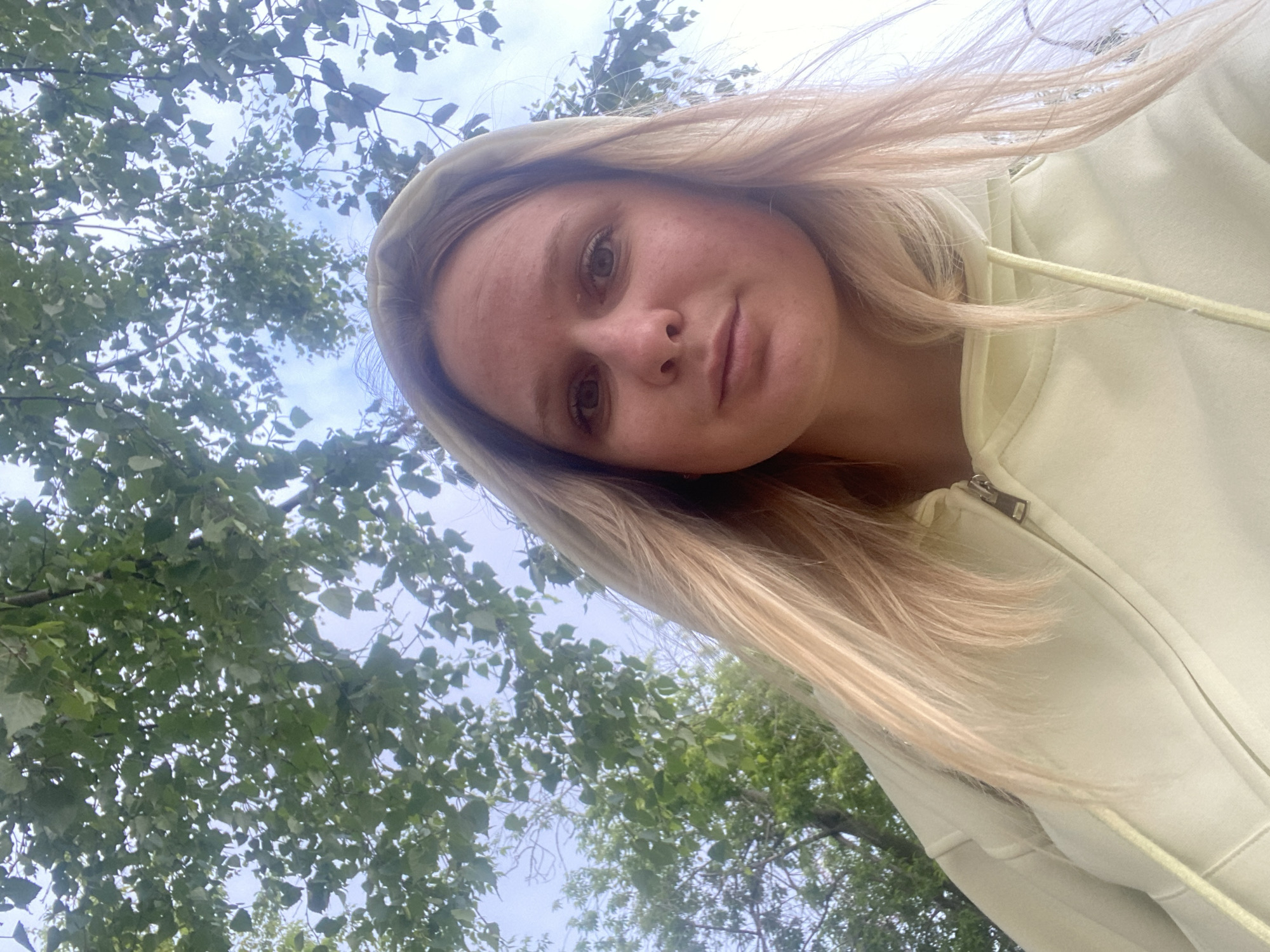 A woman with blonde hair wearing a light yellow hoodie is photographed from a low angle. The background features foliage from trees with leaves, against a partly cloudy sky. The location is unknown and cannot be determined by the contents of this image.