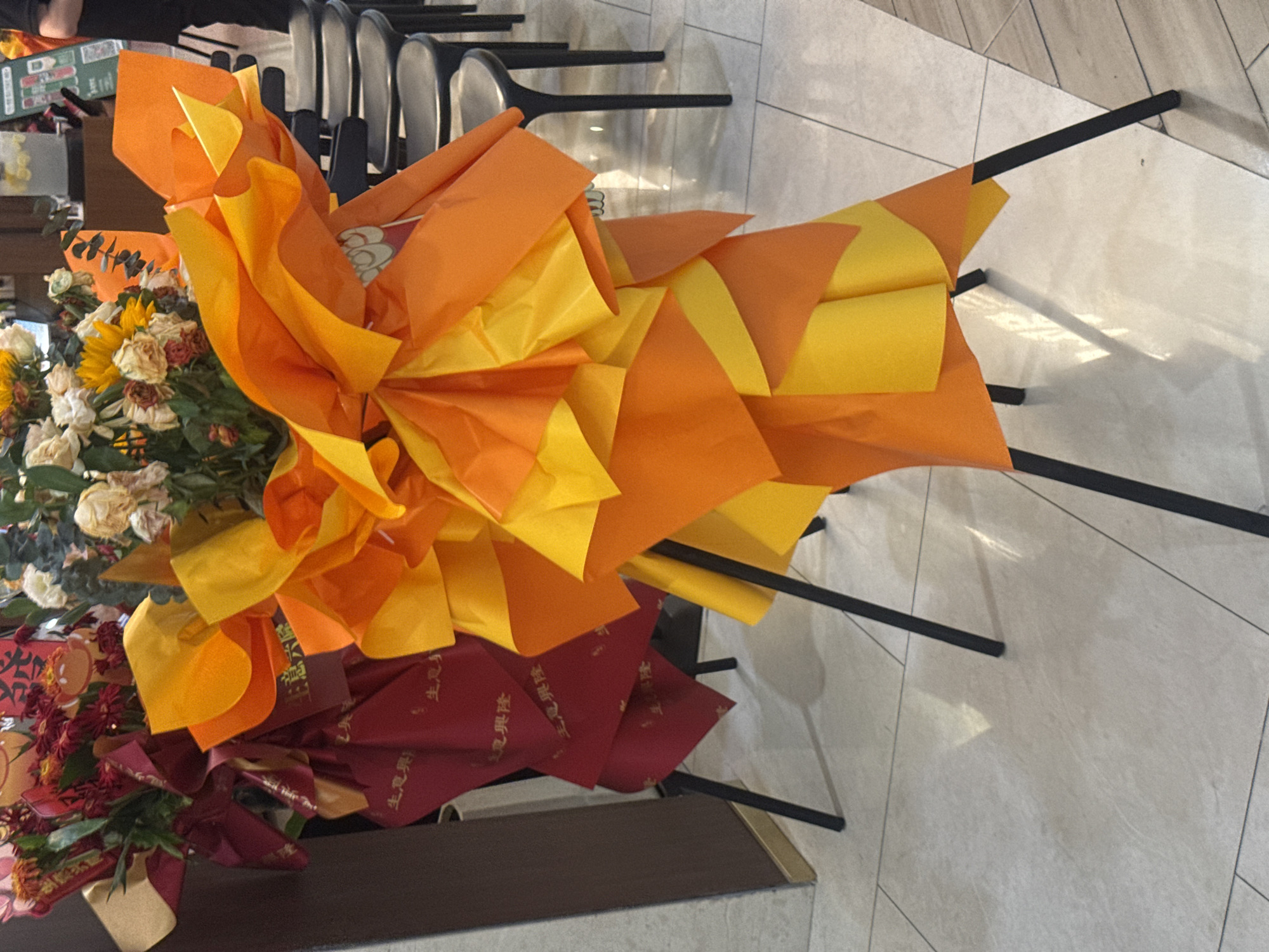 The image shows a close-up view of two elaborately decorated floral arrangements, likely for a grand opening or celebration, displayed indoors on a tiled floor. The arrangements are primarily composed of bright orange and yellow paper and flowers, creating a festive and eye-catching display. In the background, several black chairs are visible, arranged in a row, suggesting a public or commercial space. One of the floral arrangements features a red banner with gold Chinese characters, which are difficult to decipher clearly but appear to be congratulatory in nature. The lighting suggests an indoor environment, possibly a shop, event venue, or a corporate office in Qinhuangdao, China. There are no people clearly visible in the frame, focusing the attention entirely on the floral displays and the surrounding environment. The floor is made of light-colored tiles, and the overall impression is one of a celebratory event or establishment.