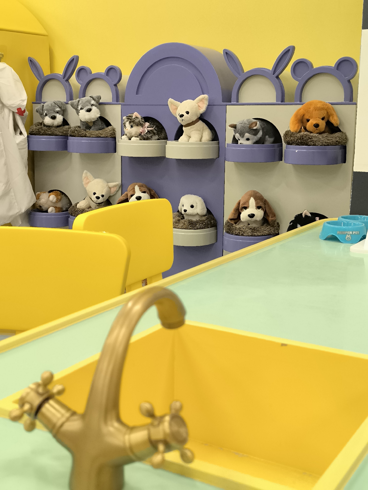 This is a photograph of a play area in the city of Qarağandı, Kazakhstan, likely a children's daycare or a play center. The focus of the image is a wall unit designed to resemble animal kennels, filled with stuffed toy dogs. The kennels are arranged in rows and feature decorative bunny and bear ears at the top. In the foreground, a yellow sink with a brass faucet is visible, suggesting the play area includes a pretend kitchen or grooming station. The overall color scheme is bright and cheerful, with yellow walls and purple accents on the kennel unit.