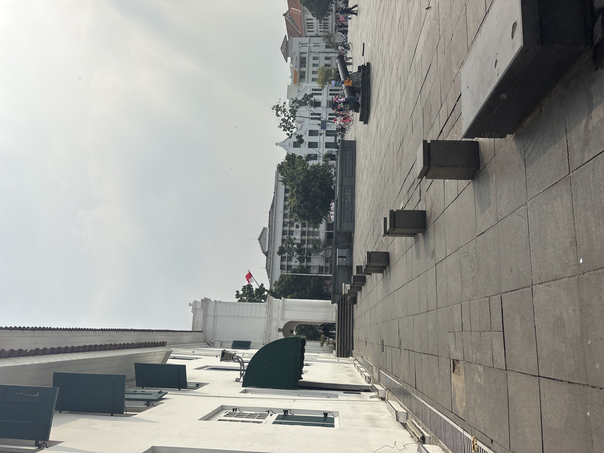 The image captures a view of Fatahillah Square in Kota Tua, West Jakarta, Indonesia. On the left side, a white colonial-era building with green shutters is visible, casting a shadow on the gray stone-tiled pavement. In the distance, several other historic buildings with red-tiled roofs and white facades are present. A prominent feature in the square is an old cannon on a stone base, with a few people around it, including children. The sky is overcast, and the lighting suggests it is daytime. Scattered throughout the square are low concrete planters and a long, dark bench. The overall scene depicts a public square with historical architecture and a few individuals engaged in casual activity.