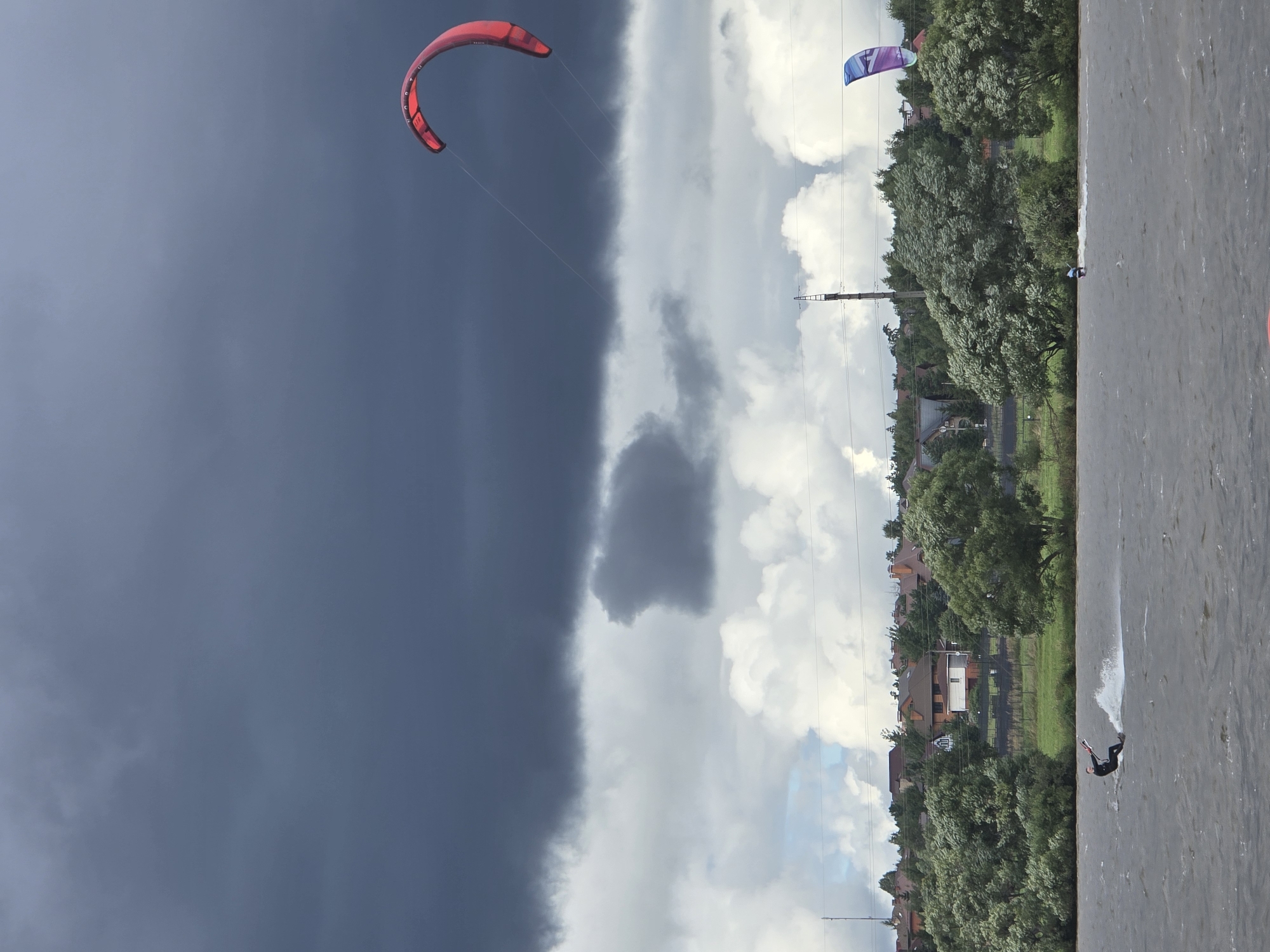 The image depicts a kitesurfing scene on a body of water, likely a lake or bay, near Novoseltsevo, Russia. The sky is dramatic, dominated by dark, heavy storm clouds in the upper portion of the frame, juxtaposed with brighter, puffier clouds lower down. In the distance, a treeline with scattered houses and buildings forms the backdrop.
In the foreground, a kitesurfer in a dark wetsuit is actively engaged in the sport, carving through the choppy water and leaving a white spray in their wake. They are holding onto a kite string, which connects to a large, red kite angled upwards in the sky. Another, smaller kite, colored purple and blue, is visible further in the distance, to the right of the frame, also with a kitesurfer beneath it. The water is a deep gray, reflecting the overcast sky. The overall impression is one of dynamic weather conditions and outdoor recreation.