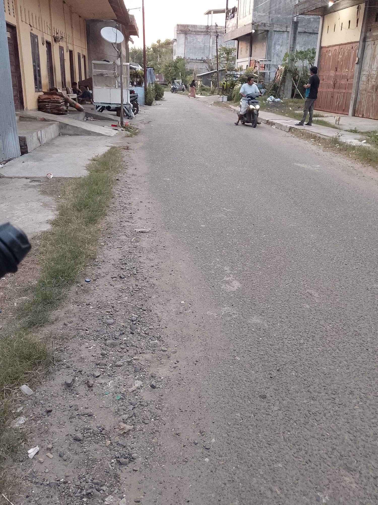 This media file depicts an everyday street scene in Lhokseumawe, Indonesia, likely during the late afternoon or early evening given the warm, soft lighting. An asphalt road dominates the center, stretching into the distance. It is bordered by dirt and gravel shoulders with sparse green grass, and raised concrete sidewalks on both sides.
On the left side of the street, a multi-story building with multiple doors is visible. In front of it, two individuals are seated on a low platform or bench, near stacked cardboard boxes. A white mobile vendor cart with a glass display is parked nearby, surmounted by a satellite dish on a pole. Another motorcycle is parked behind this cart. Further down the street, a person is seen walking away from the camera on the left.
On the right side of the road, various concrete buildings with large, reddish-brown gates line the street. A man in a light-colored shirt rides a black scooter on the asphalt, moving away from the camera. Ahead of him, on the sidewalk, another man in a dark shirt and pants stands, holding what appears to be a bottle or bag. A small, light-colored cat walks on the sidewalk near his feet.
The scene conveys a sense of local urban life with residents going about their day. Some small pieces of litter, including plastic and a blue bottle cap, are scattered on the ground near the grass. No visible text is discernible in the image.
